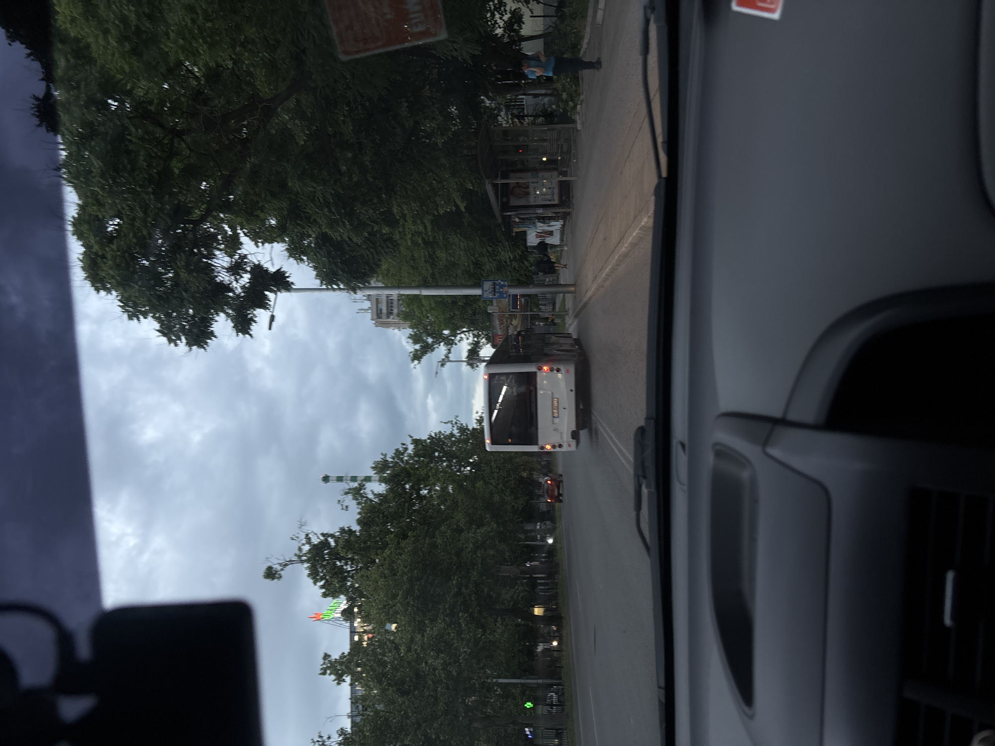 The image shows a view from inside a vehicle, likely a car or van, looking out onto a street in Plovdiv, Bulgaria. A white bus is directly ahead, traveling in the same direction. The road is lined with trees and sidewalks. Pedestrians are visible near a bus stop on the right side of the road. In the distance, towards the lower portion of the frame, a large green and white structure, possibly related to an amusement park or fair, is visible. Overcast skies are present.