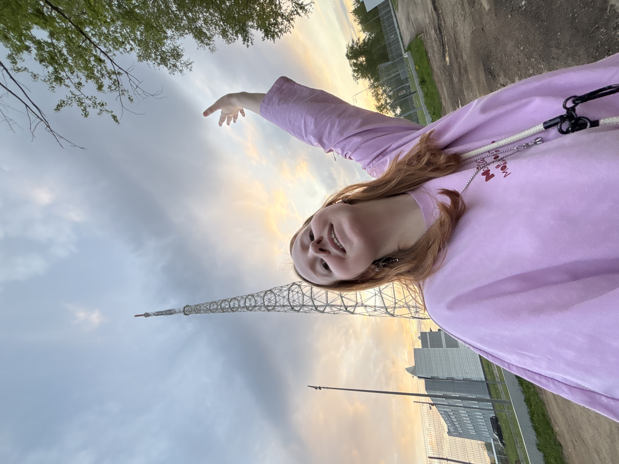 The image captures a rotated selfie of a young person, likely female, with light brown hair, smiling broadly. She is wearing a light purple long-sleeved top. Her left arm is extended upwards and to the left, with her index finger pointing, as if gesturing towards something or simply posing.
The scene is outdoors during the golden hour, either sunrise or sunset, indicated by the warm orange, pink, and yellow hues of the sky near the horizon, which transitions to a cooler grey-blue higher up. The sky is partially cloudy.
Prominently in the background is a tall, lattice-style radio or television tower, consistent with the Perm TV Tower in Perm, Russia, given the provided context. Below the tower and to the right, several modern, multi-story buildings are visible, suggesting an urban or industrial setting. Green foliage from tree branches with leaves is present in the upper left corner of the frame. A chain-link fence and a dirt path or road are also visible towards the right side of the image.
The person appears happy, indicated by her wide smile. A silver chain necklace with a small pendant rests on her chest. On her purple top, below the necklace, some dark red text is partially visible, appearing to spell "MooN". A white rope-like strap with black connectors, possibly from a bag, is seen over her right shoulder.