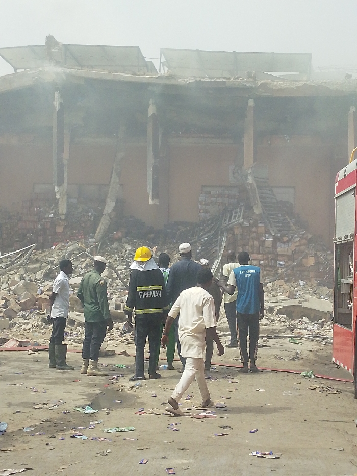 The image depicts a post-disaster scene in Fagge C, Nigeria, showing a multi-story commercial or storage building that has sustained severe structural damage, likely from a fire. The upper floor and roof have partially collapsed, leaving exposed beams and concrete slabs, with solar panels askew on the remaining roof structure. A substantial mound of rubble, including shattered concrete, bricks, and numerous stacked boxes, has accumulated at the base and within the lower levels of the building. Heavy smoke or dust haze permeates the air around the structure.
Approximately ten adult males are observed in the foreground and midground, surveying the scene. A firefighter, centrally positioned and facing the damaged building, wears a yellow helmet and a dark uniform with "FIREMAN" emblazoned on the back. To the firefighter's left, two men stand, one in a white shirt and dark trousers, the other in a green uniform. To the right, civilians in various attire, including one wearing a white skullcap and another in a blue t-shirt marked "LFC UNION," are present. In the immediate foreground, a man in a light-colored traditional tunic walks away from the damaged building. A red fire truck is partially visible on the far right, with red hoses extended across the dusty ground, which is littered with small, colorful wrappers and debris.