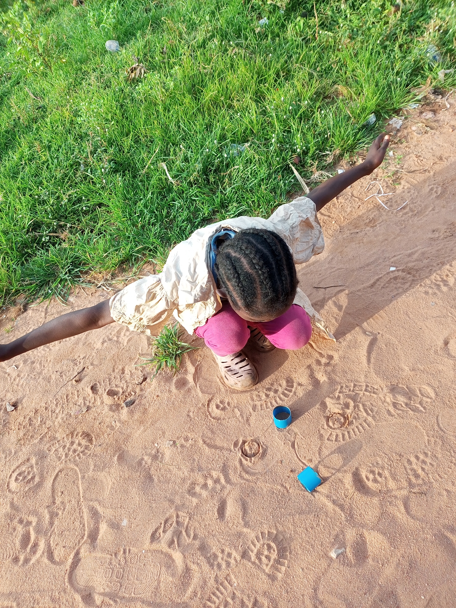 In Tempure, Nigeria, a young Black girl, with braided hair, is playing in the sand. She is wearing a cream-colored dress and pink leggings. Her arms are outstretched, and she is looking down at the sand. There are two blue cylindrical objects and several footprints in the sand around her. A patch of green grass borders the sandy area.