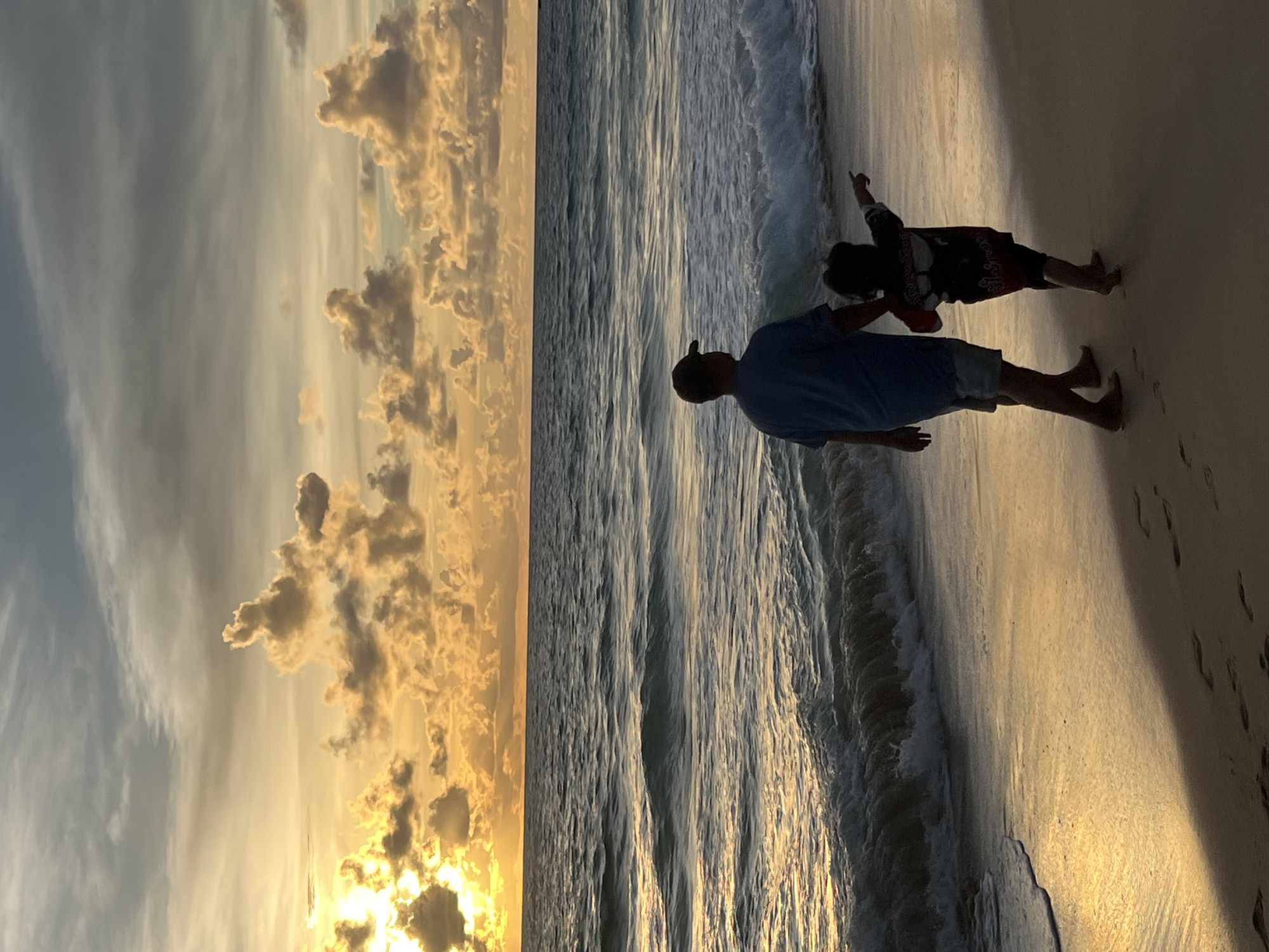A father and child walk hand-in-hand along a sandy beach at sunset in Choeng Thale, Thailand. The child, wearing a dark t-shirt with red accents, points towards the ocean. The father, in a blue t-shirt and shorts, wears a baseball cap. Gentle waves lap at the shore, reflecting the golden hues of the setting sun. The sky is a canvas of dramatic clouds, illuminated by the sun's warm light. Footprints mark the wet sand, hinting at their leisurely stroll. The scene captures a serene moment of connection amidst the natural beauty of a tropical coastline.