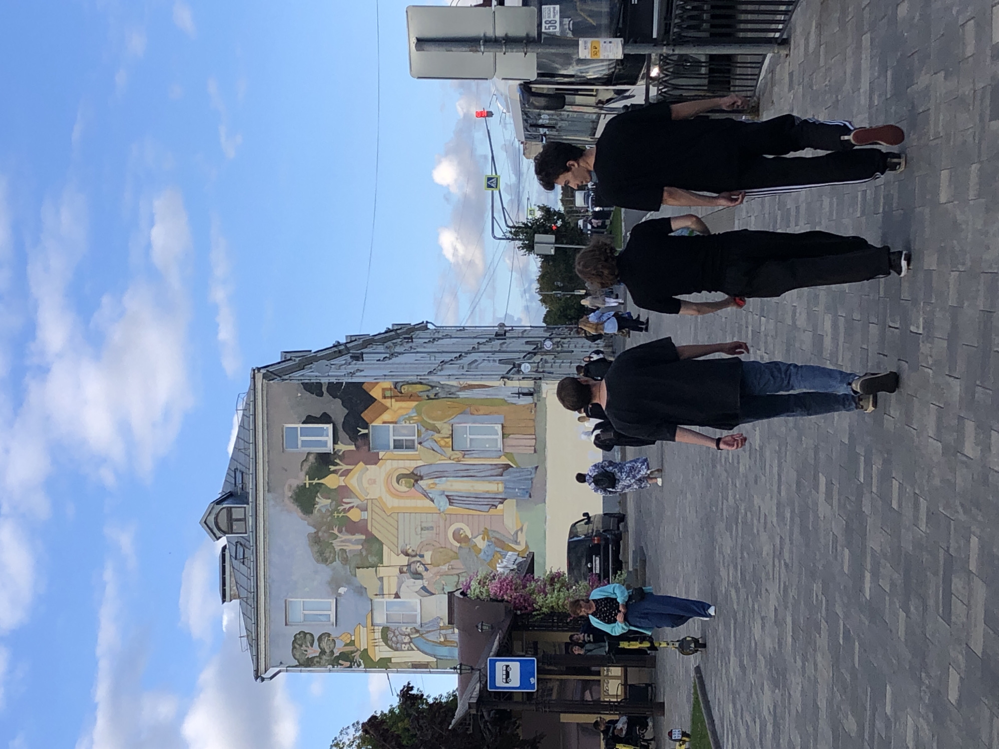 The image depicts a street scene in Sergiyev Posad, Russia, with a large mural covering the side of a building. Several people are walking on a paved sidewalk. In the foreground, three young men are walking away from the camera. The man on the left is wearing a black t-shirt and blue jeans. The middle man is wearing a black t-shirt and black pants. The man on the right is wearing a black t-shirt and black pants with white stripes.
Further down the sidewalk, a woman in a blue cardigan and dark pants is walking towards the camera, with a bus stop shelter to her left. Behind her, other pedestrians are visible, some carrying backpacks. The building on the right has a mural that appears to depict religious figures and scenes, with typical Russian Orthodox architectural elements like onion domes. The sky is blue with scattered white clouds. A bus is visible on the street to the right, and traffic lights and street signs are present. The overall atmosphere suggests a normal day in a public urban space.