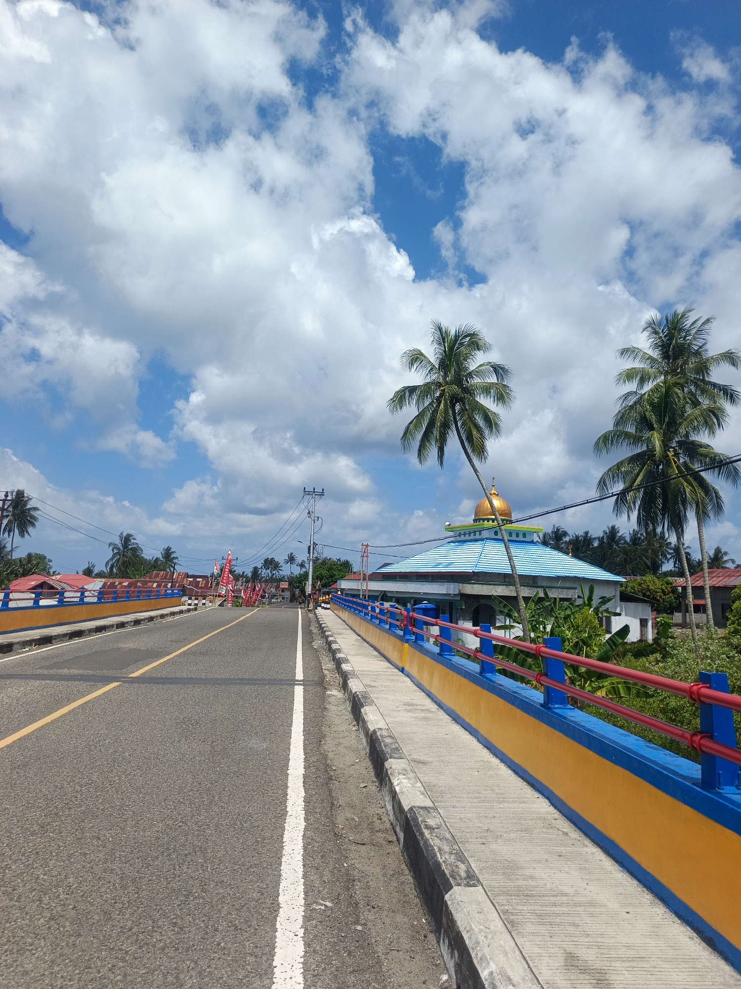 This is a vertical, eye-level shot looking down a bridge in Sogitia, Indonesia. The bridge has a bright yellow base with blue railings and red poles. The road is asphalt with a yellow line down the center. On the right side of the bridge is a sidewalk. In the distance on the right, there is a building with a light blue roof and a golden dome, likely a mosque, surrounded by lush green vegetation and palm trees. On the left side of the bridge, there are several buildings with red roofs and flags visible. The sky is a vibrant blue filled with fluffy white clouds. The overall scene depicts a sunny day in a tropical environment.
