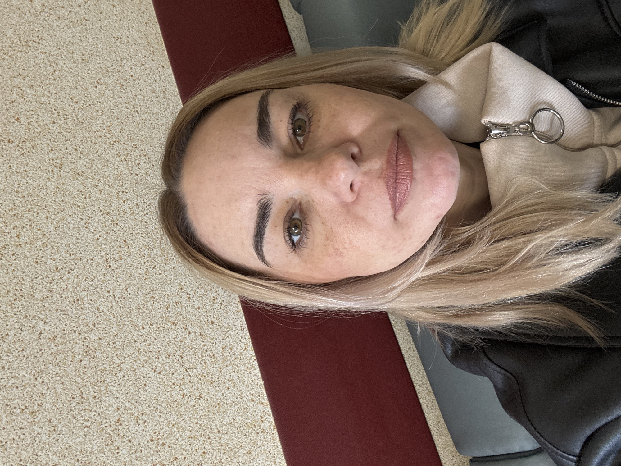 A woman with blonde hair and green eyes is looking directly at the camera. She has freckles and defined eyebrows. She is wearing a black leather jacket with a beige hoodie underneath. The background includes a speckled beige surface, a dark red vertical stripe, and a gray surface. There is no evidence indicating this photo was taken in Saint Petersburg, Russia.