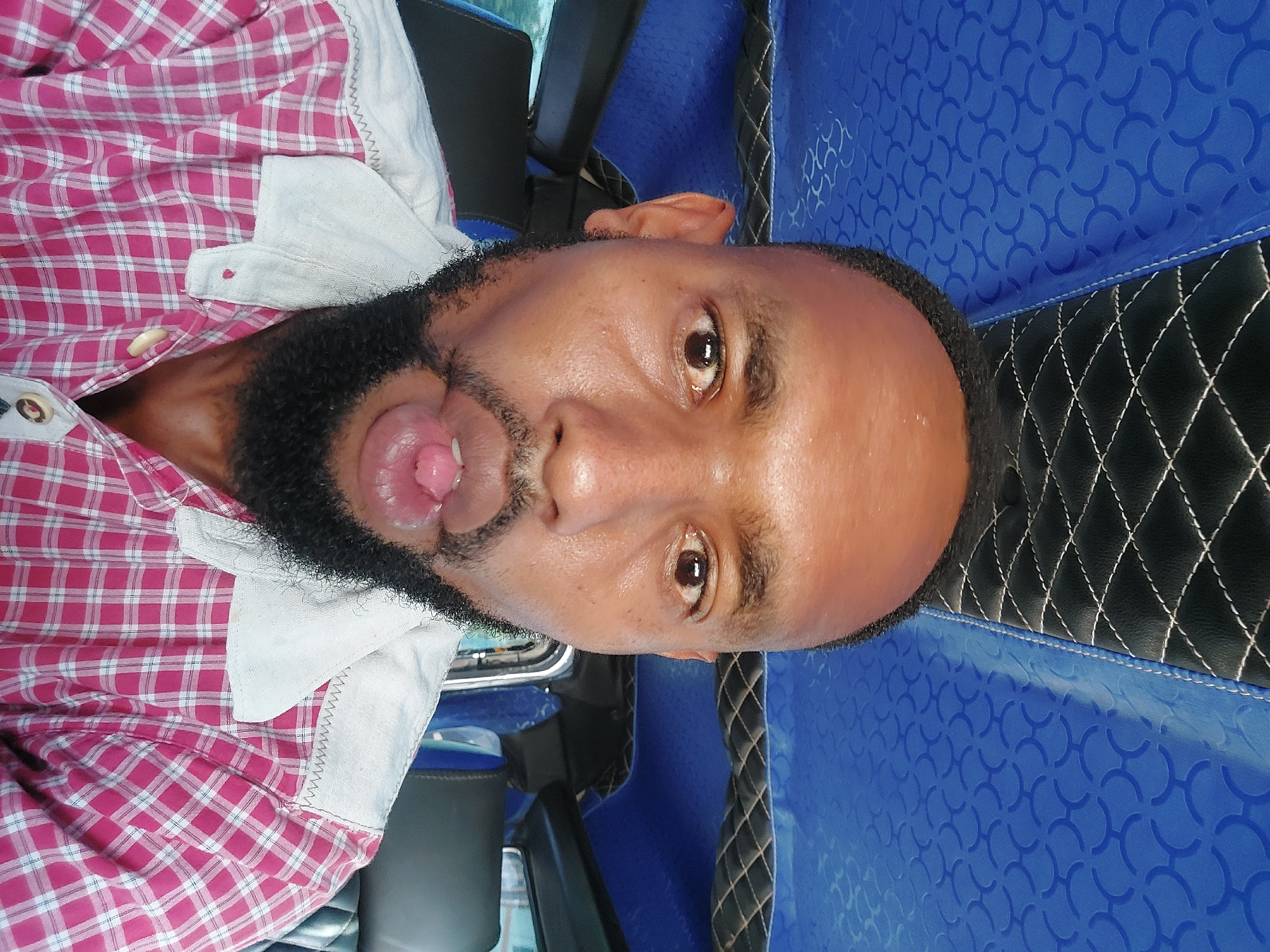 A close-up, slightly angled headshot depicts an African male with dark skin, a closely shaved head, and a short, dark beard. The man is wearing a pink and white checkered shirt with a white collar. His mouth is open, revealing his tongue. The background features the interior of a vehicle, identifiable by the blue patterned material and black stitched leather. It is not possible to confirm location in Dar es-Salaam, Tanzania from the image.