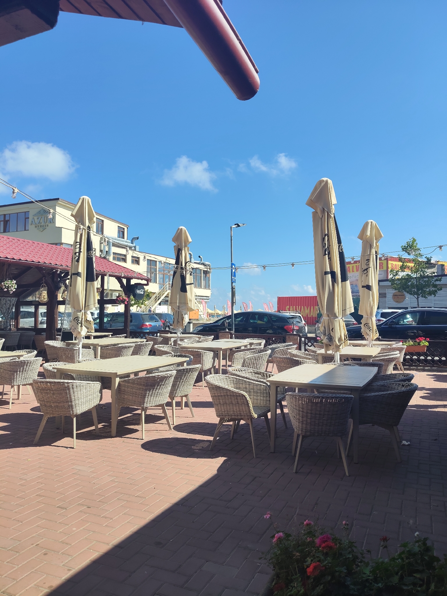 The image shows an outdoor seating area of a restaurant or cafe, likely located in Constanța, Romania, on a sunny day. The foreground is dominated by rows of outdoor tables and chairs made of wicker, arranged on a brick-paved patio. Several large, closed umbrellas are positioned between the tables, casting shadows on the ground. In the background, a multi-story building with a red-tiled roof is visible, featuring the "AZUR EVENTS" logo. Beyond this building, more structures and parked cars are present, suggesting an urban or semi-urban setting. The sky is clear blue with a few scattered clouds. The overall impression is that of a pleasant, open-air dining space.
