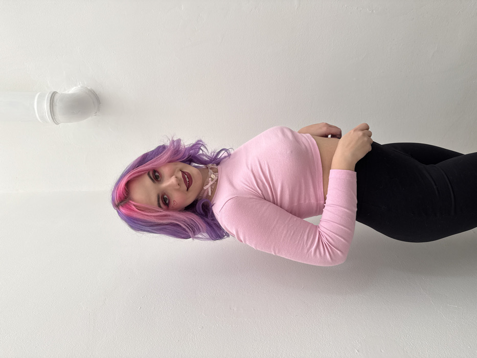 A young woman with purple and pink hair poses against a white background. She is wearing a light pink long-sleeved crop top and black leggings. A white pipe fixture is visible in the upper left corner of the frame. The woman is smiling and wearing a ribbon choker necklace. A small heart is drawn on her cheek. She appears to be leaning against the wall at an angle.