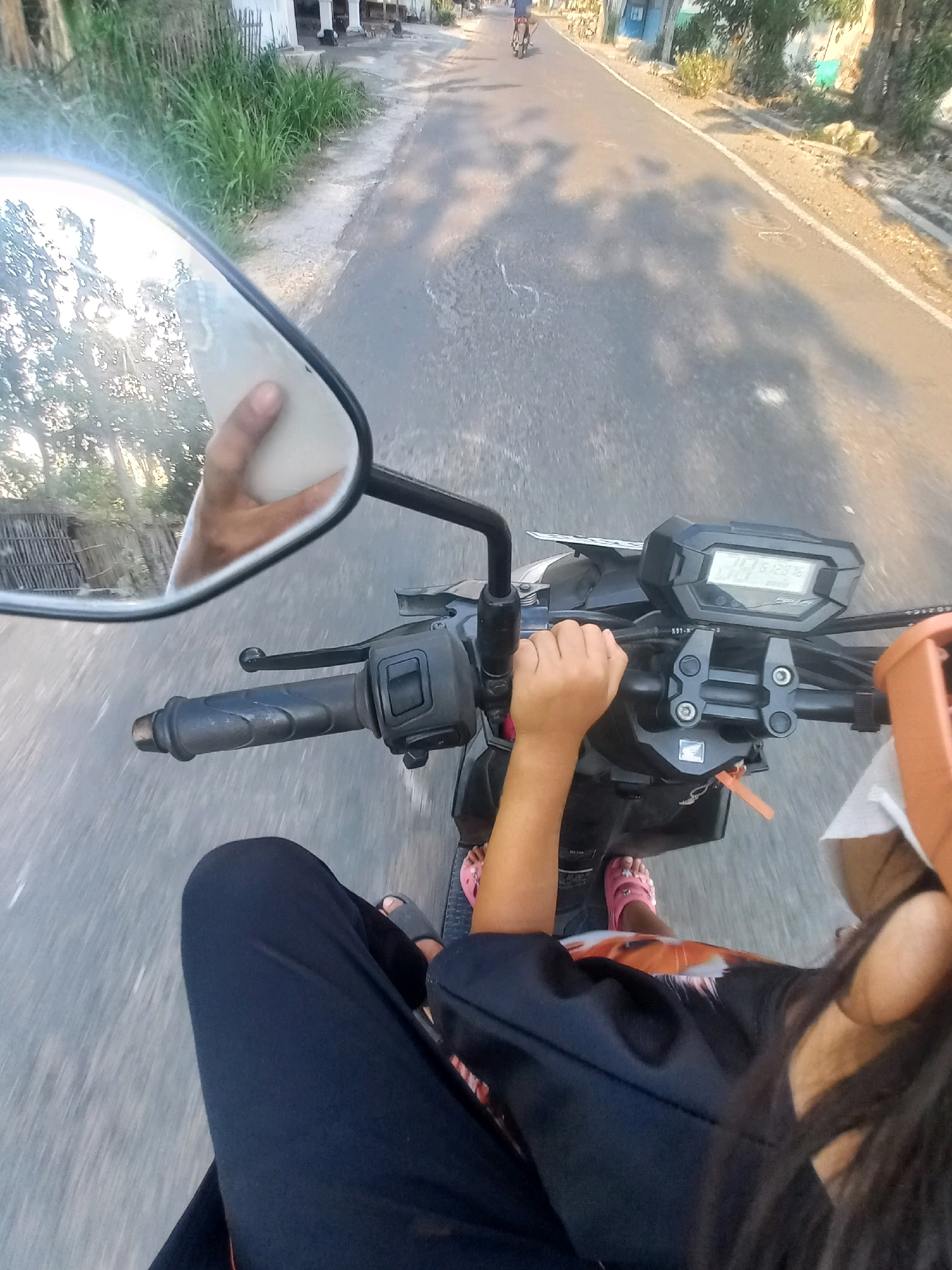 The image depicts a first-person perspective of someone riding a motorcycle on a paved road in Baturetno, Indonesia. The rider's hands are visible on the handlebars, and their legs are extended forward. The motorcycle has a digital display panel. In the foreground, the rider's black pants and pink sandals are visible. The background shows a road with sparse vegetation and buildings along the sides. The sun is casting shadows on the road, indicating it is likely daytime. The overall impression is a typical scene of transportation within a semi-urban or rural Indonesian setting.