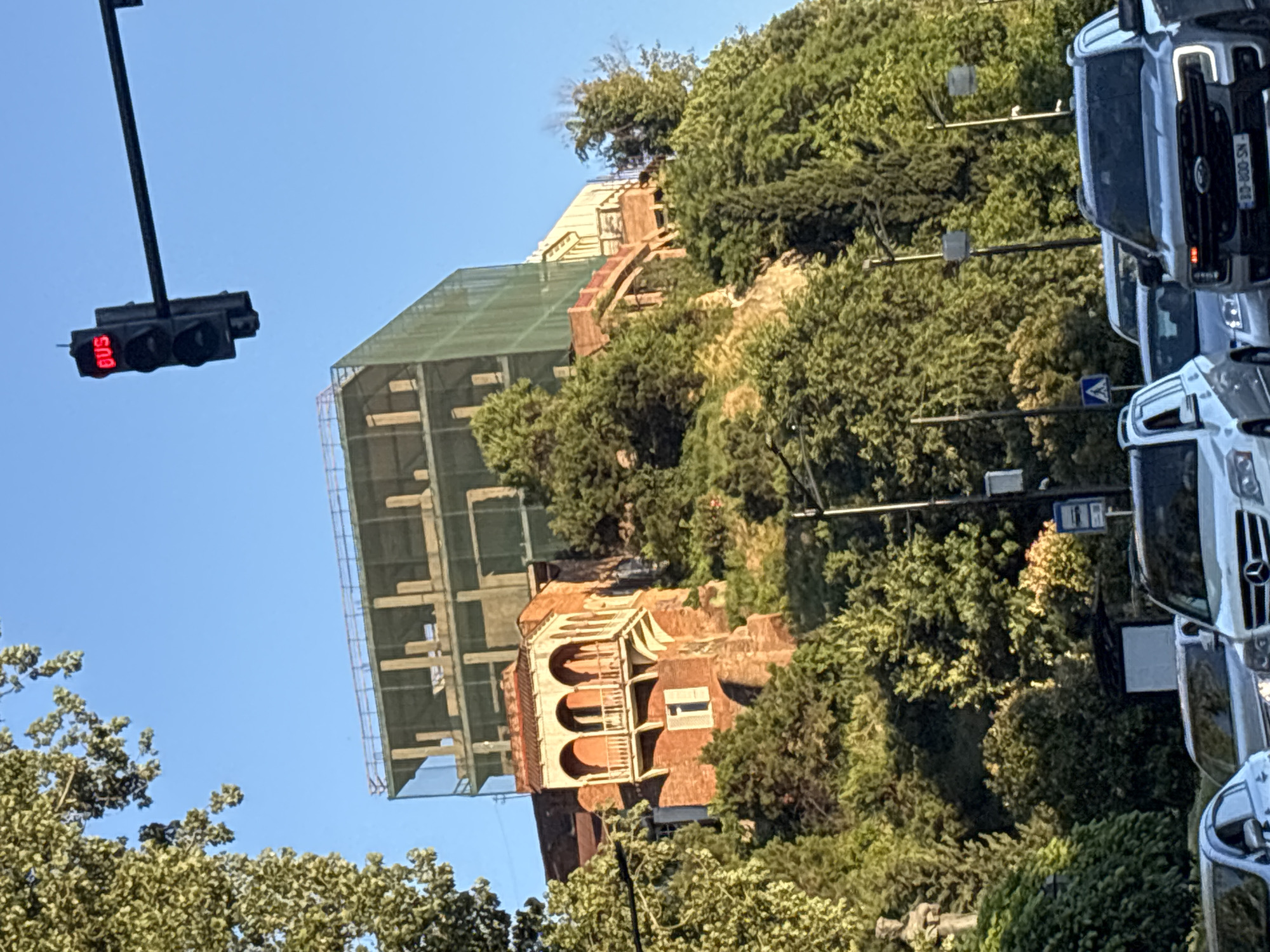 The image shows a multi-story building under construction, partially covered with green netting, situated on a steep, tree-covered hillside in Tbilisi, Georgia. In the foreground, several cars are visible on a road, indicating an urban setting. A traffic light with a red "BUS" display is positioned in the upper right quadrant of the image. The architecture of the older, lower section of the building appears to be of brick with arched windows and balconies, contrasting with the modern construction above. The hillside is densely vegetated with various trees. The sky is clear and blue. The overall scene depicts a city undergoing development, with historical structures alongside new construction.