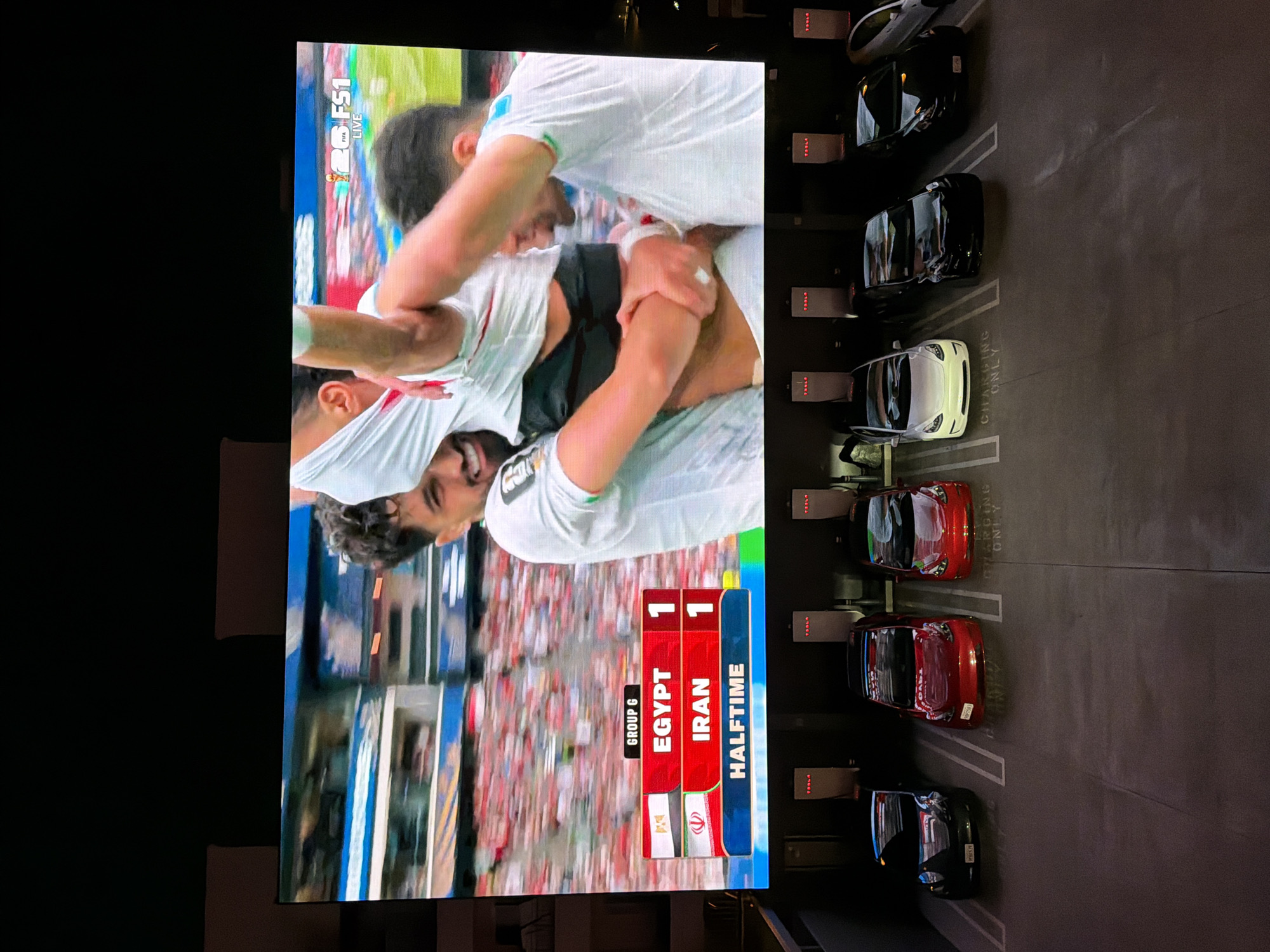 In West Hollywood, California, a large outdoor LED screen displays a live broadcast of a soccer match between Egypt and Iran, with the score at halftime being 1-1. The screen is positioned above a row of Tesla charging stations, with several electric vehicles parked and charging. The charging stations are labeled with the Tesla logo and are integrated into the parking lot infrastructure. The overall scene suggests a public space or commercial area where attendees can watch sporting events while their vehicles are being charged.