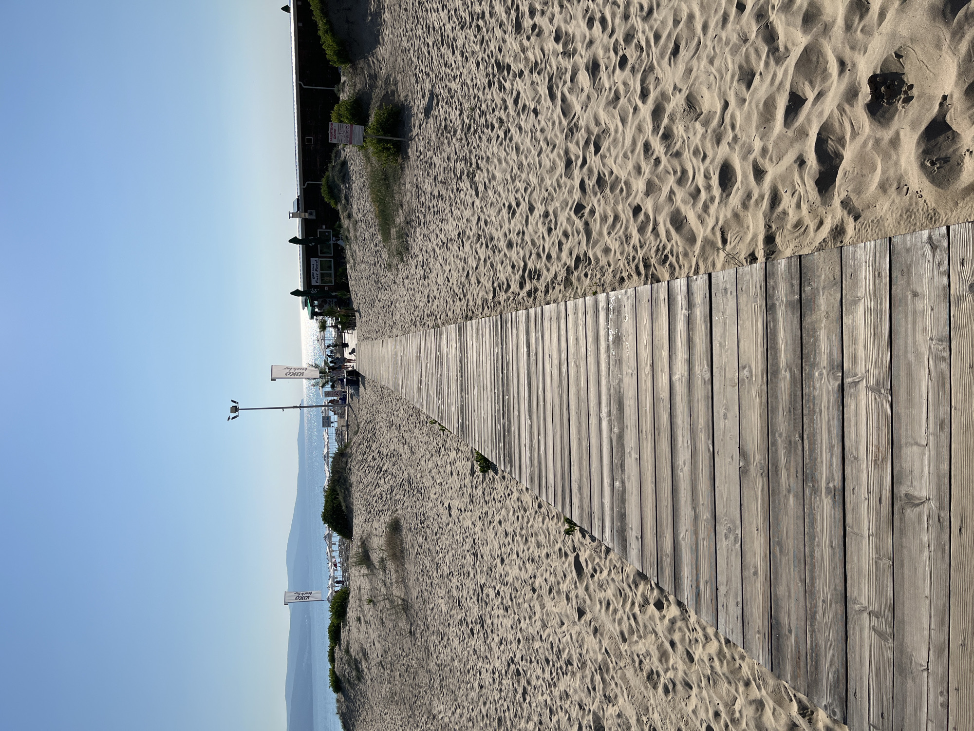 A wooden boardwalk stretches across sandy beach terrain toward the sea under a clear blue sky. The boardwalk, constructed of weathered grey planks, is the dominant foreground element, leading the viewer's eye towards the horizon. To the left and right of the boardwalk, the beach is composed of fine, light-colored sand, sculpted by wind into rippled patterns. Sparse patches of dry vegetation dot the sand dunes.
In the middle ground, the boardwalk approaches a low-lying building situated on the beach. This building, with a dark facade and a white roof, appears to be a beach bar or restaurant, as suggested by umbrellas and tables visible in front of it. Flags, one white with blue lettering that reads "UKO beach bar", are planted in the sand near the building. A lamppost stands to the right of the boardwalk. A few individuals are visible in the distance, walking along the boardwalk and near the building.
The background features the calm, sparkling blue sea, reflecting the sunlight. Distant hills or mountains are visible on the horizon, under a vast, cloudless sky. The overall impression is a serene beach scene on a bright, sunny day. This scene is located on the beach in the resort town of Sunny Beach, near Nesebar, Bulgaria.