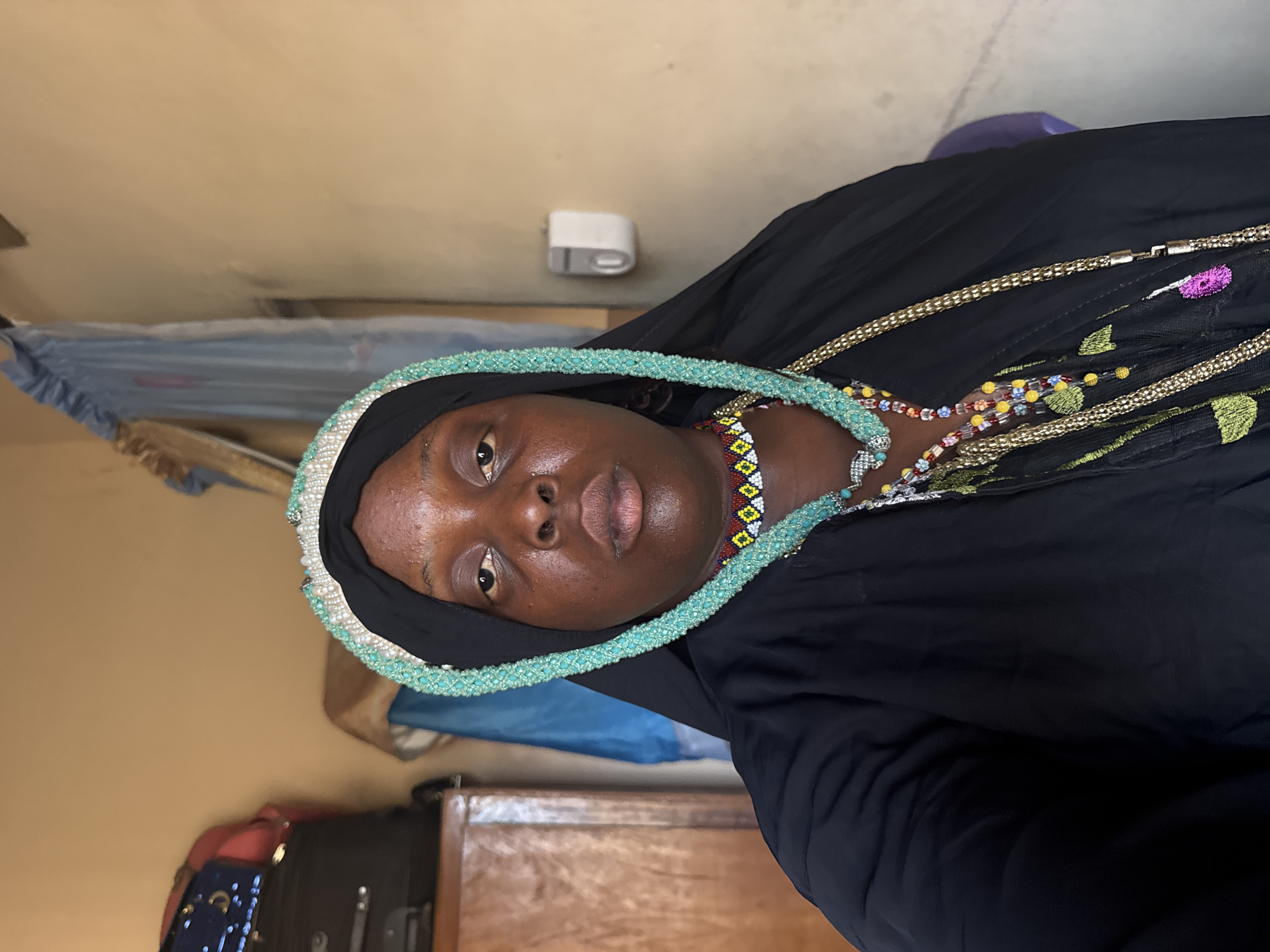 A medium shot features a woman with dark skin wearing a black hijab. A turquoise beaded necklace is around her neck along with a multicolored beaded necklace, a gold chain, and a black embroidered shawl. The room has beige walls with a white electrical device mounted high and a bed partially visible in the background. There's also a dark wooden dresser and suitcase at the bottom of the frame. The location is Madangala, Nigeria.