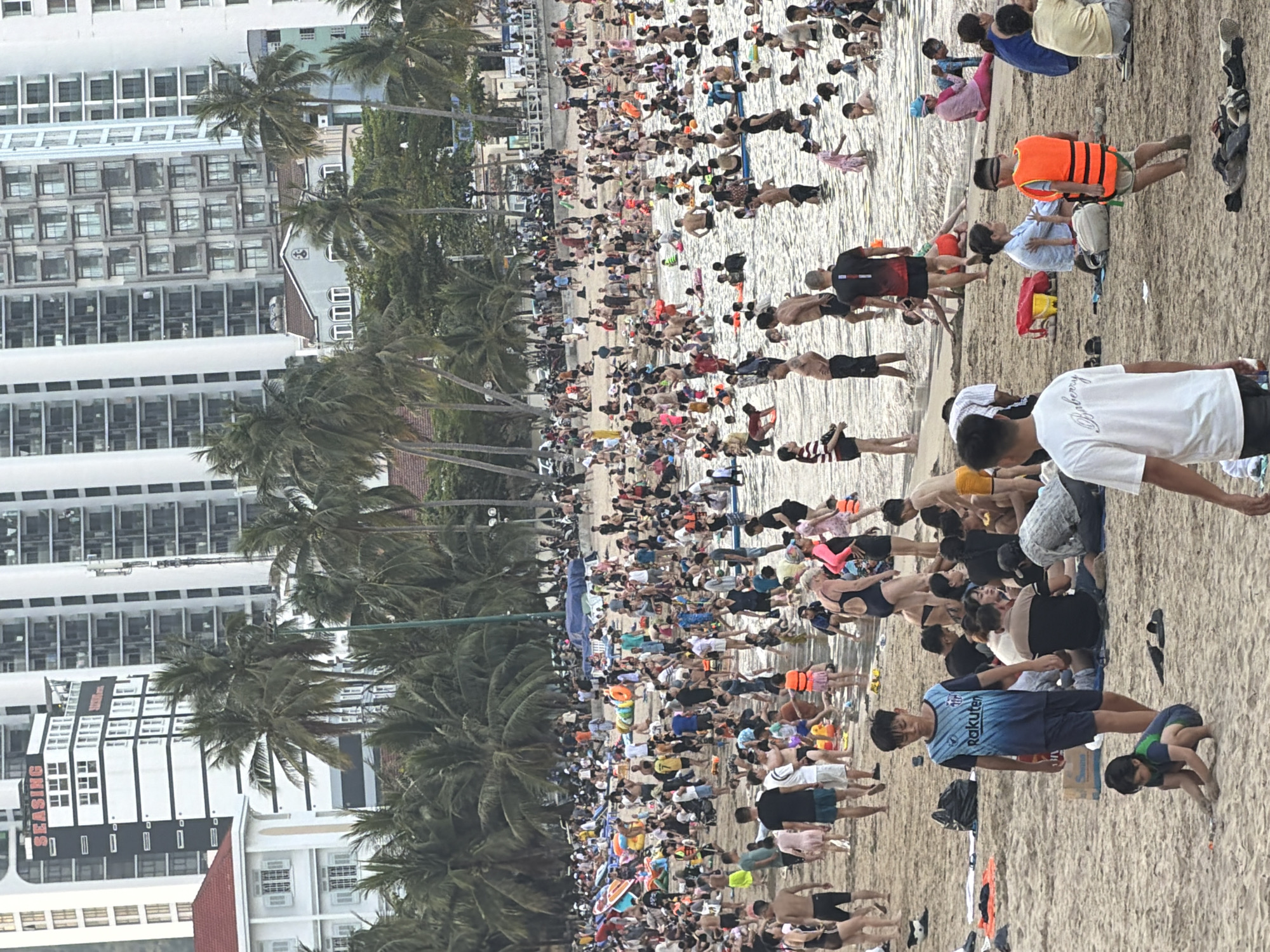 This image depicts a crowded beach scene in the city of Nha Trang, Vietnam, likely during daylight hours. The background features numerous high-rise buildings and palm trees, characteristic of a coastal urban environment. The foreground and midground are populated by a large number of people, varying in age and attire, engaged in various beach activities. Some individuals are wading in the shallow water, while others are relaxing on the sand. There are numerous inflatable toys and personal flotation devices visible, suggesting recreational water activities. Several people are wearing swimwear, while others are in casual clothing. The sandy beach is partially covered by towels and bags. The overall impression is one of a busy public beach during a time of high activity.