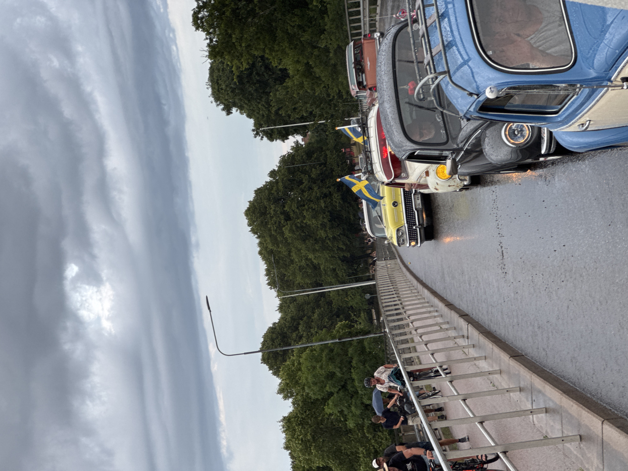 A convoy of vintage cars, several adorned with Swedish flags, drives down a wet bridge in Falkenberg, Sweden. The sky is overcast, suggesting rain. People on bicycles are visible on the sidewalk to the left of the cars. The cars include a yellow classic car, a red station wagon, a black Volkswagen Beetle, and a blue and cream Volkswagen Beetle in the foreground. The bridge has a metal railing along the pedestrian walkway. Trees line the background.