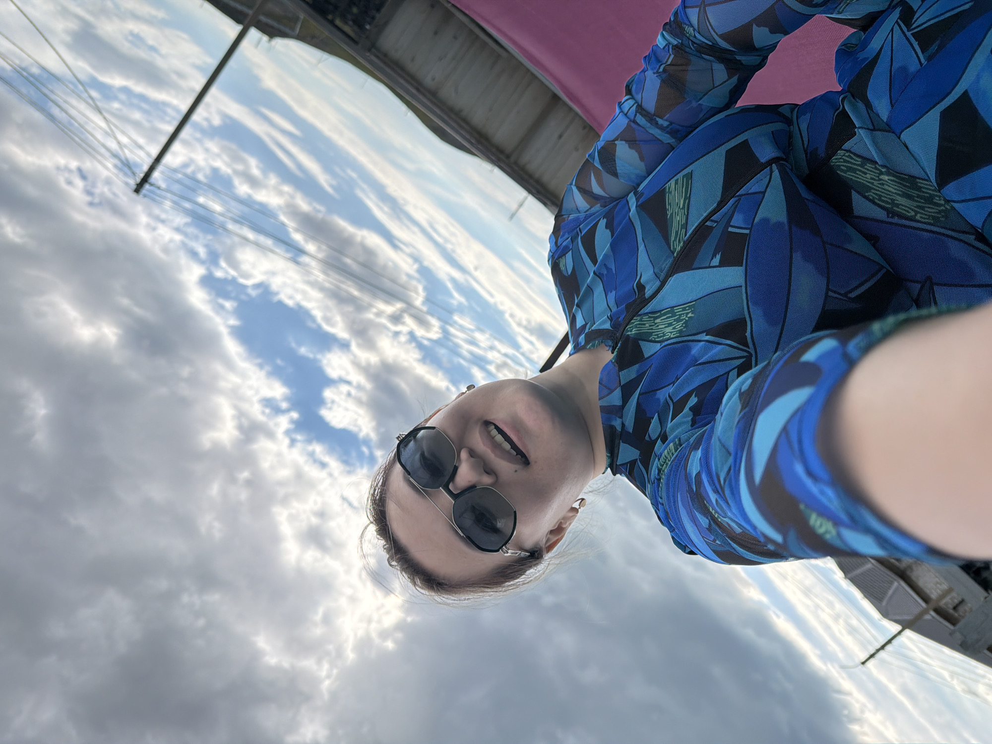 A low-angle, close-up selfie features a woman with brown hair pulled back, wearing dark sunglasses and a blue patterned mesh dress. She is smiling broadly, with her mouth open. Behind her, the sky is filled with dramatic clouds, with some streaks of blue visible. A utility pole with several wires stretches diagonally across the frame from the upper right to the mid-left. In the background, a dark, sloped roof structure, possibly part of a building, is visible. To the right of the woman, a bright pink material, possibly fabric or upholstery, creates a contrasting backdrop. The overall impression is of an outdoor setting, possibly on a rooftop or balcony, in Молбаза, Kazakhstan, under a cloudy sky.