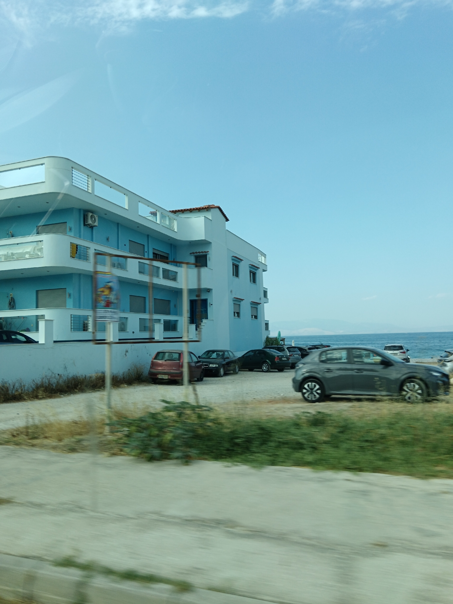 The image depicts a blue building located in the Municipal Unit of Sikyona, Greece, situated near the sea. The building has multiple levels with balconies and windows. In front of the building, a parking area is visible with several cars parked, including a gray hatchback, a red hatchback, and a black sedan. The sea is present in the background, with a hazy landscape visible on the horizon. The sky is clear with some wispy clouds. The foreground shows a paved area and some vegetation, suggesting a roadside or public space. The image appears to be taken from a moving vehicle, as indicated by motion blur in the foreground.