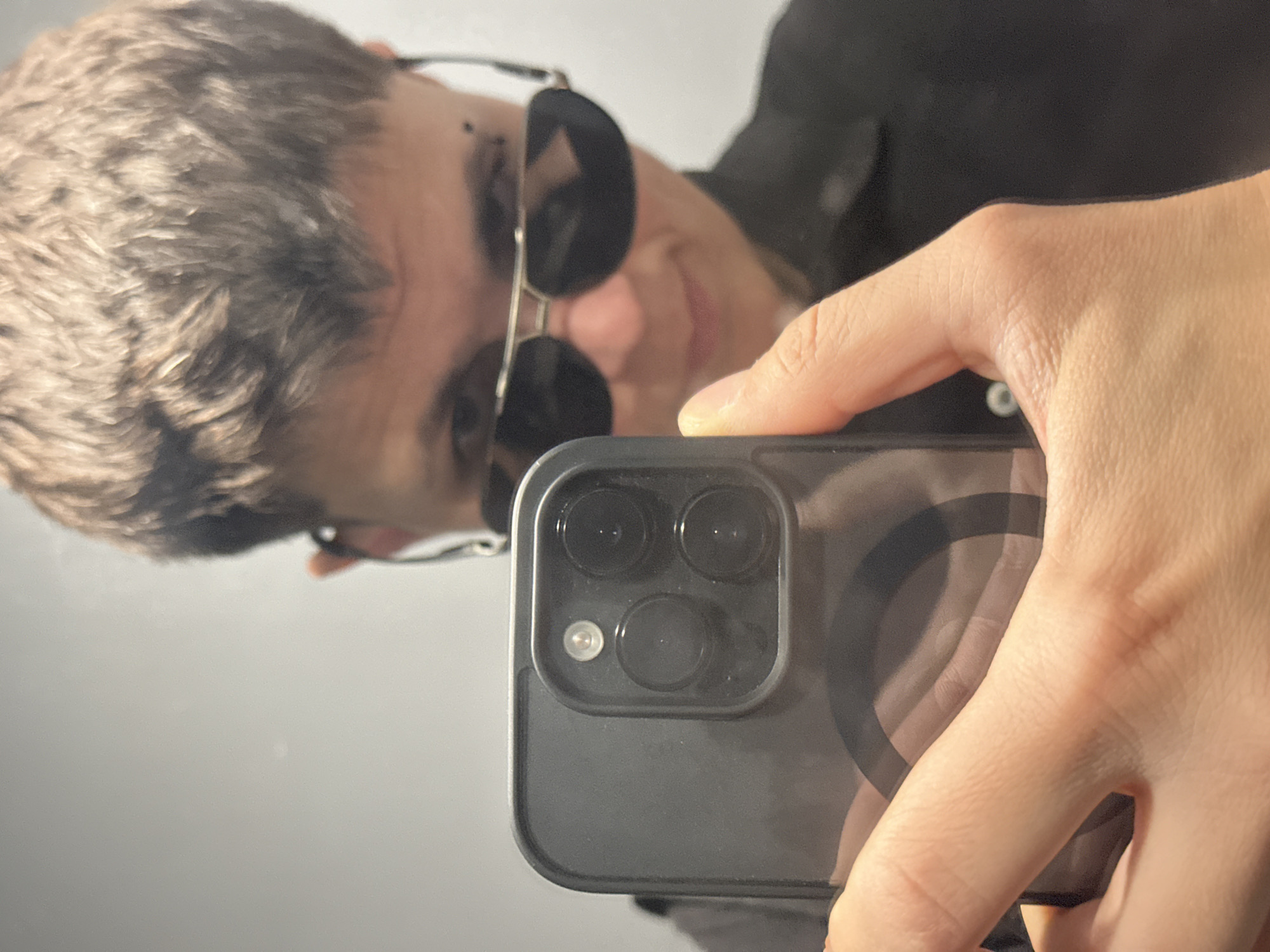 A person is taking a selfie with a smartphone in a room. The person has short brown hair and is wearing black sunglasses and a black shirt. The smartphone has a triple camera lens system. The background is a plain gray wall. There is no visible indication of the location in the city of Uzhhorod, Ukraine.