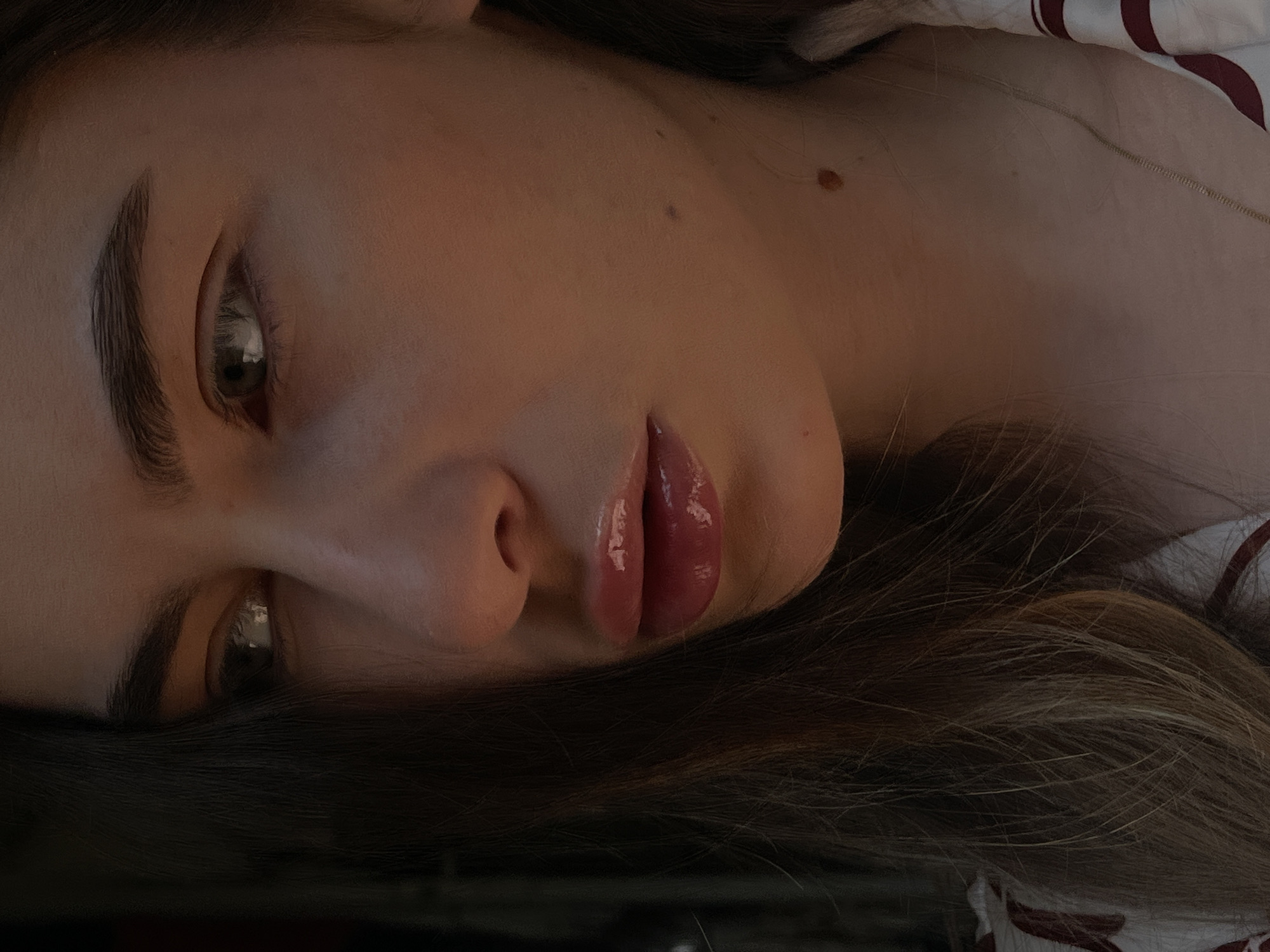 A close-up image depicts a person with fair skin lying down. The individual has noticeable eyebrows, eyes that appear to be gazing towards the left of the frame, and lips with a glossy finish. The person's hair, dark brown with lighter highlights, is visible beneath their face. Patches of bare skin are visible. The subject appears to be resting on a striped bedsheet, with the stripes in shades of white and dark red.