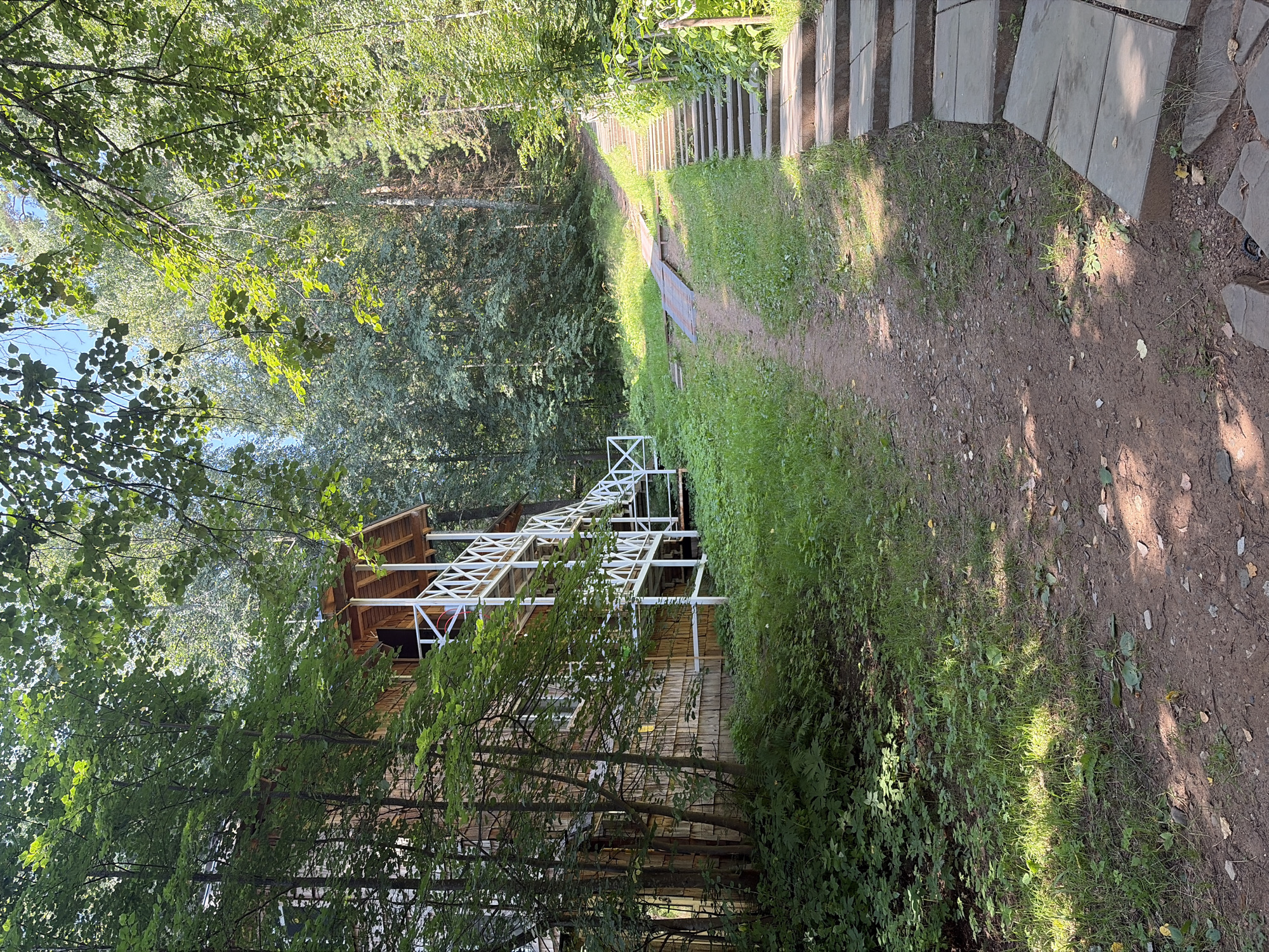 The image depicts a wooden building with a white, multi-level external staircase and railing, nestled amongst dense green foliage in a forest setting. The building appears to be a house or cabin with a rustic wooden exterior. A dirt path with some stone slabs leads away from the building, winding through the grass and trees. Sunlight filters through the leaves, casting dappled shadows on the path and ground, suggesting it is a sunny day. The overall atmosphere is peaceful and natural. The location is identified as Kostovaty, Russia. There is no visible text in the image.