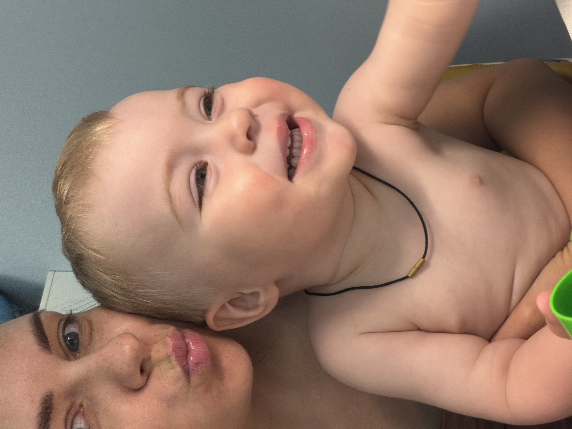 A close-up, high-angle shot captures a baby lying on their back, partially on top of a woman. The baby, who is fair-skinned with light blonde hair, is smiling with their mouth open, revealing some teeth. They are wearing a black string necklace with a gold-colored bead. The woman, presumably the mother, is seen with part of her face visible, including her eye, nose, and lips. The background is a light blue, likely a wall. The specific location in Votkinsk, Russia, is not discernible from the image.