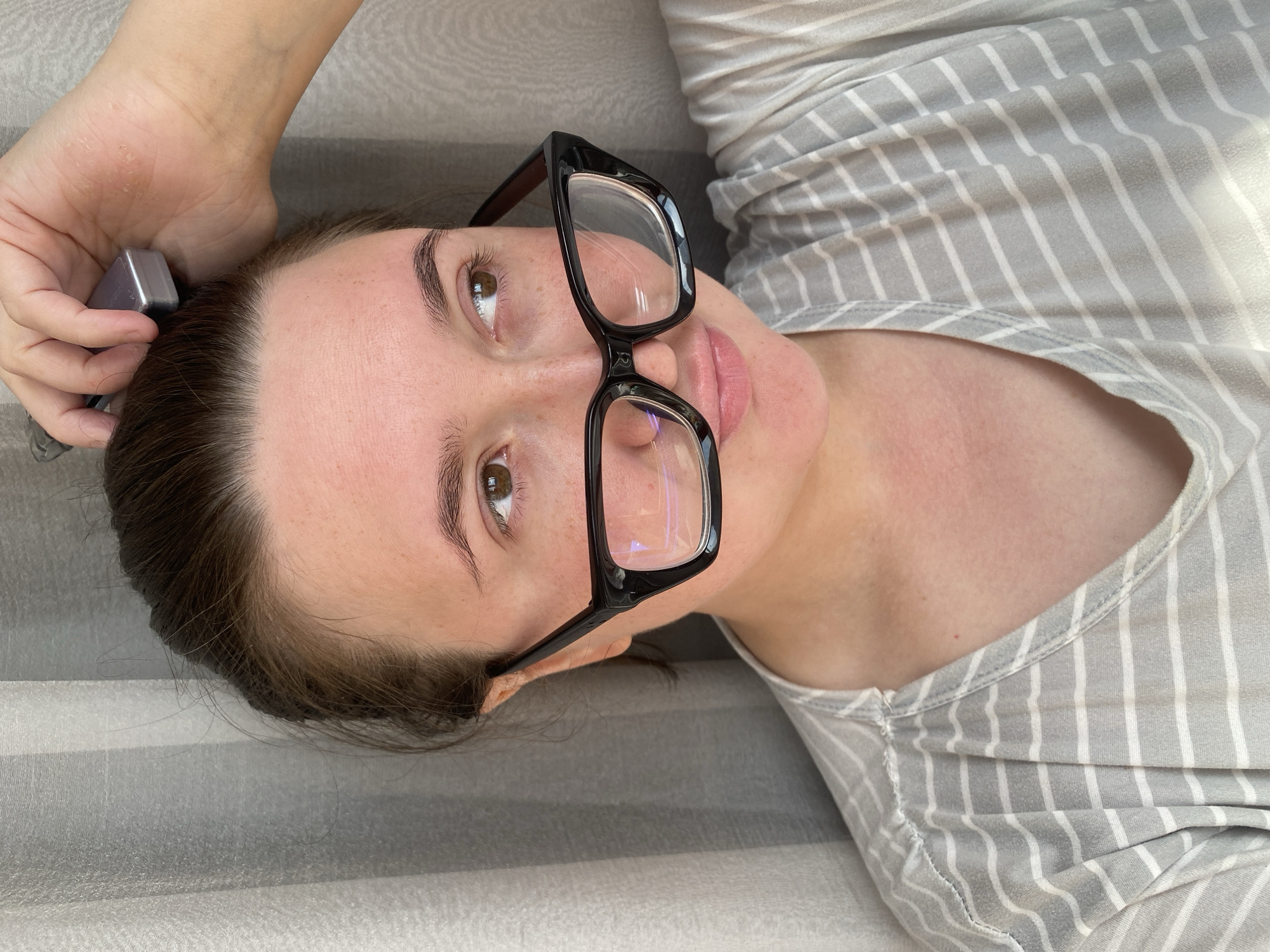 The media file features a close-up, high-angle shot of a woman lying down, taken from a perspective looking down at her. She has fair skin, dark brown hair, and dark eyebrows. Her light brown eyes are open and directed upwards. She is wearing black-framed glasses that rest on her nose. Her lips are slightly parted, revealing a neutral or contemplative expression.
The woman is dressed in a light grey top with thin white vertical stripes. Her left hand is raised towards her head, holding a small, silver-grey metallic object near her hairline. The background consists of a light grey surface with wider, subtly textured horizontal stripes, possibly a bedspread, blanket, or upholstered furniture. The lighting appears soft and natural, suggesting daytime. No discernible text is visible in the image. The setting is an indoor environment, likely within Chaikovsky, Russia, as per the provided context.