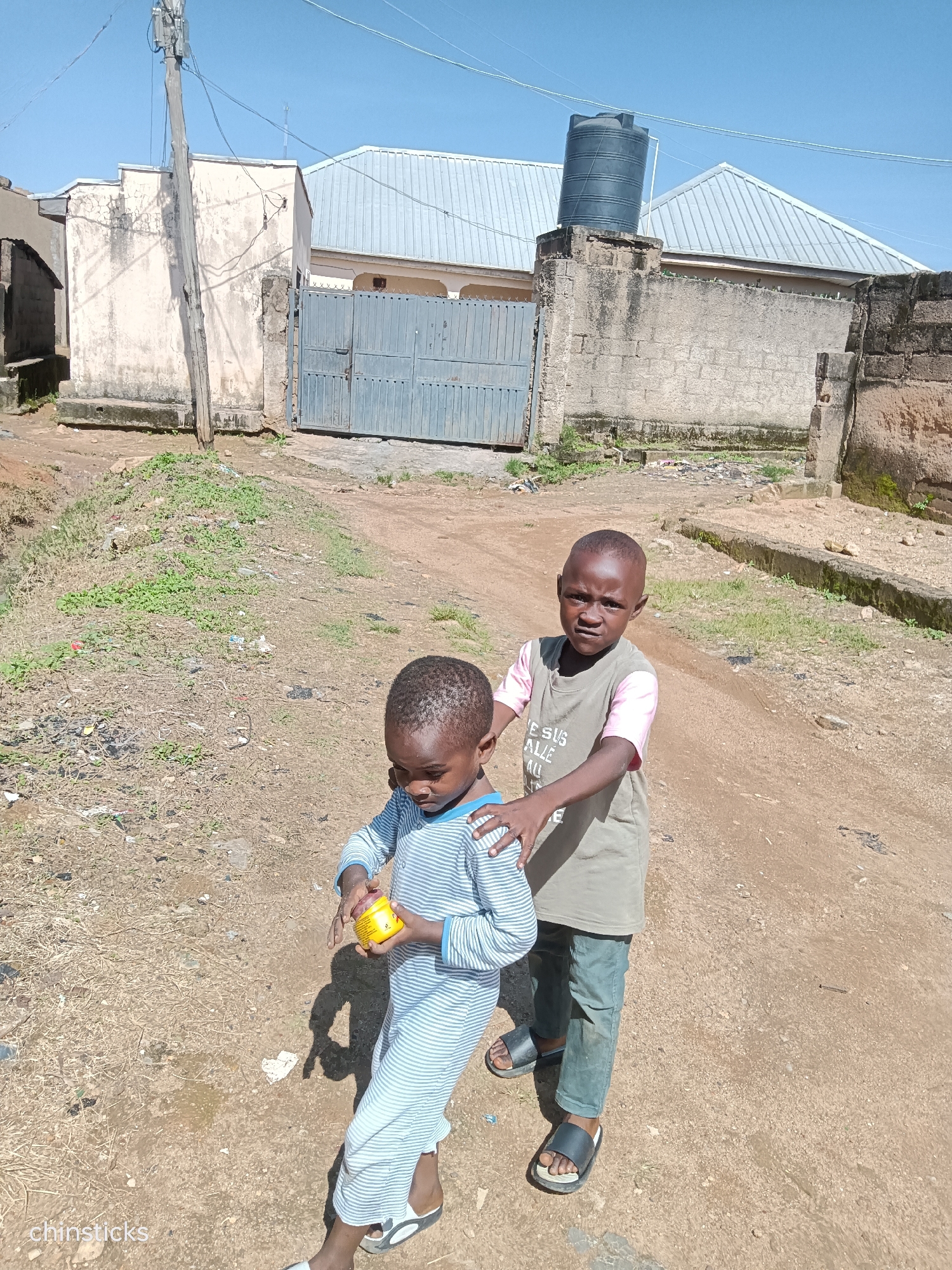 In a dirt alley in Jos, Nigeria, two young boys stand. The boy on the left wears a blue and white striped pajama set and holds a small yellow container. The boy on the right, wearing a gray t-shirt with pink sleeves and dark green pants, places his hand on the shoulder of the boy in front of him. They are in front of a building with a large metal gate and a water tank on the roof. The ground is covered in dirt and sparse vegetation, with some litter visible. The sky is clear and blue.