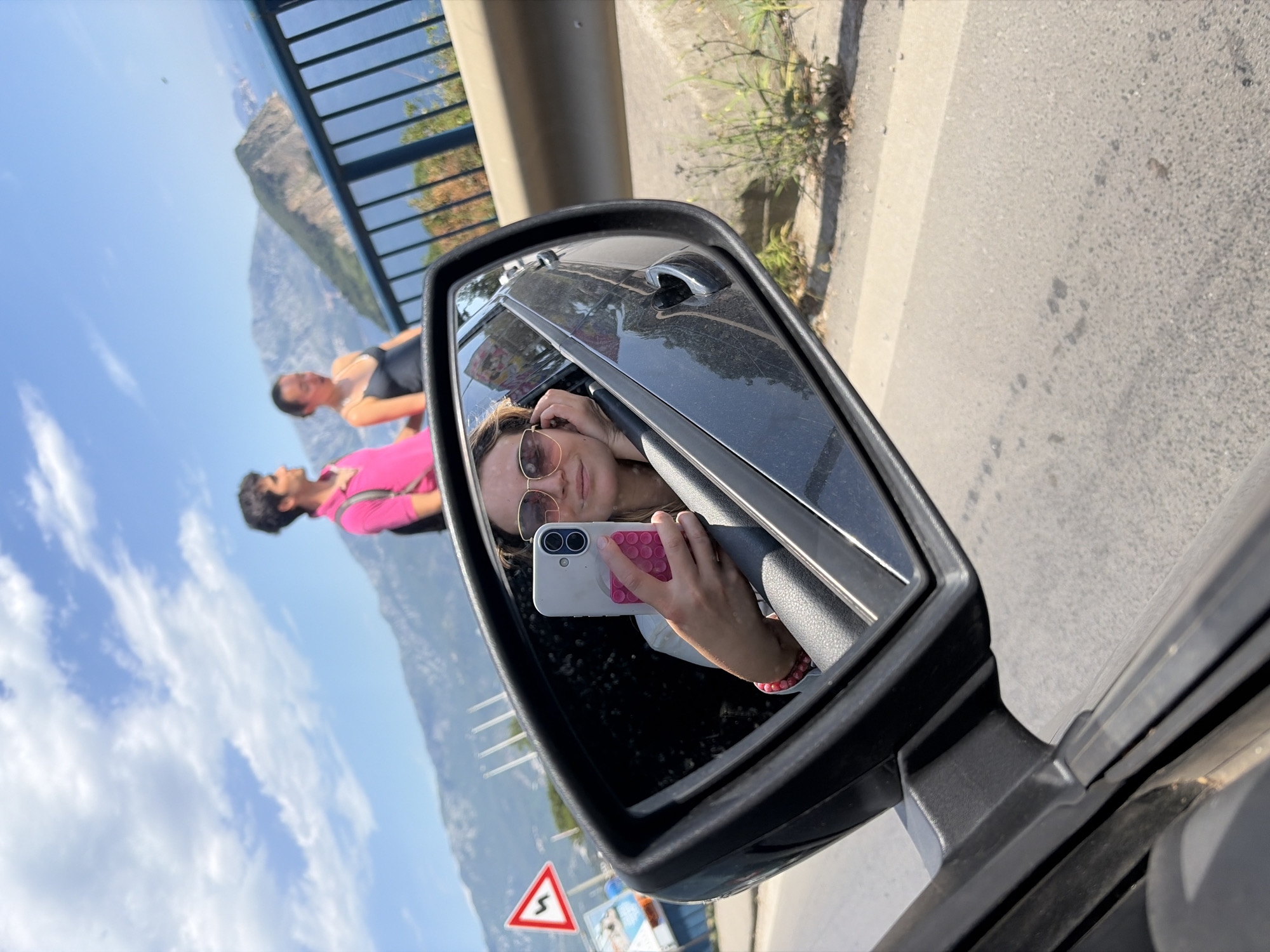 The image shows a car's side mirror reflecting a scene of Budva, Montenegro. Inside the car, a woman with sunglasses is holding a white smartphone with a pink grip, taking a picture or video. Reflected in the mirror, outside the car, are two people: a man in a pink shirt and a woman in a black top, standing near a metal railing overlooking a mountainous coastline. The sky is blue with scattered clouds. A road sign with a red border and a winding road symbol is visible in the lower part of the mirror, along with a glimpse of the road and some vegetation. The car is parked on the side of the road.