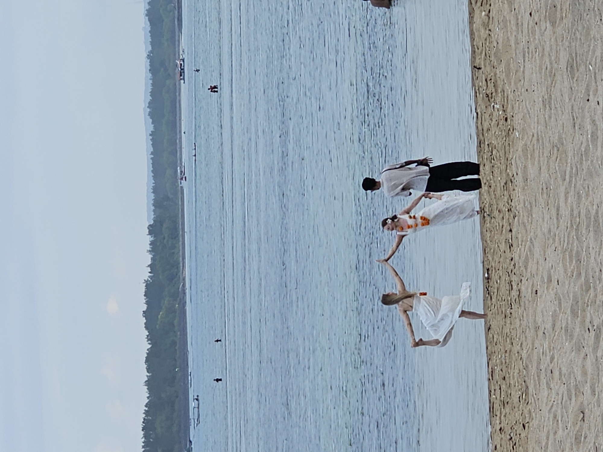 In Denpasar, Indonesia, three individuals are standing near the water's edge on a sandy beach. Two women, dressed in white, appear to be performing a yoga pose or dance, with one woman extending her arms upwards and the other in a balancing pose with her foot lifted. They are both adorned with leis of orange flowers. A man, wearing a light-colored shirt and dark pants, stands between them and slightly behind, holding hands with one of the women. The ocean stretches out before them, with several boats visible in the distance and a tree-lined shore beyond. The sky is overcast.