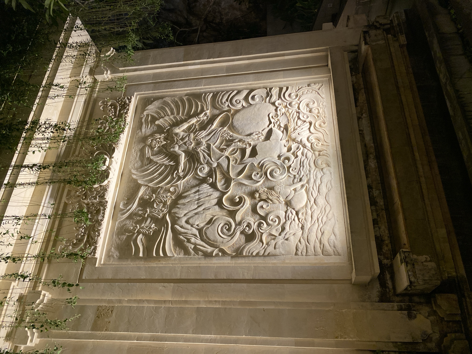 This is a nighttime photograph of a carved stone relief on a wall in Kuta Selatan, Indonesia. The relief depicts a winged figure in the center, possibly a deity or mythical creature, with mountainous terrain and swirling clouds or water in the background. The carving is illuminated by embedded lights, casting shadows that accentuate the detail. Above the relief, decorative stonework is visible, and hanging green vines cascade down from the top of the structure. The wall itself is made of stone blocks, with vertical fluting on the left side. In front of the wall are several stone steps. The overall impression is of an outdoor architectural feature, likely part of a temple or a well-decorated building. The dark background suggests it is evening or night.