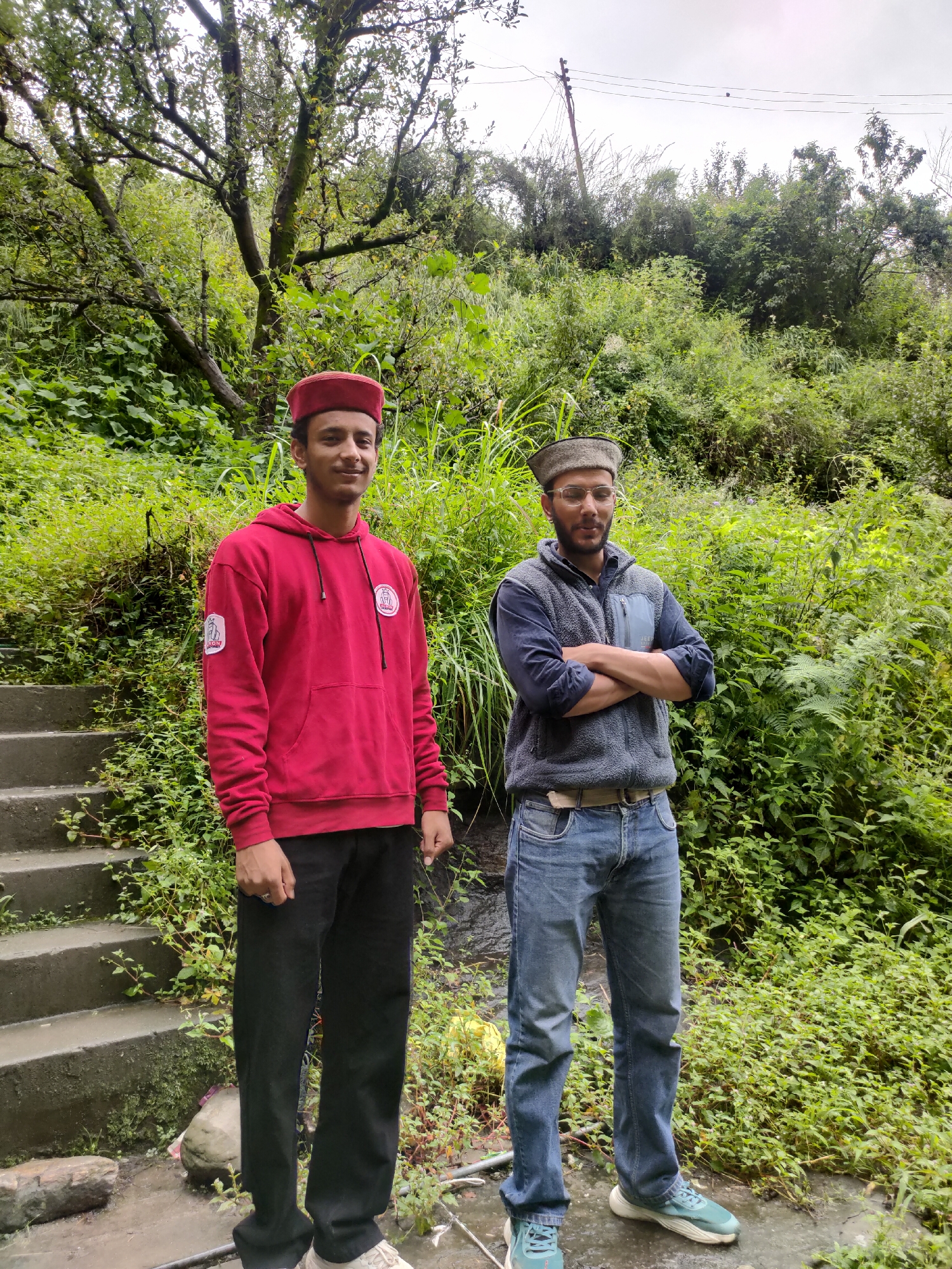Two men are standing in a lush, green outdoor environment. The man on the left is wearing a red hooded sweatshirt, black pants, and a red cap. He is smiling and facing the camera. The man on the right is wearing a gray fleece vest over a dark blue shirt, blue jeans, and glasses. He has his arms crossed and is looking towards the right side of the frame. They are standing on a paved surface with concrete steps to the left and dense vegetation surrounding them. The background consists of trees and overgrown plants, suggesting a natural or semi-rural setting. A power line pole and wires are visible in the upper right corner of the image. The presence of stairs and a paved path suggests this may be part of a trail or a residential area in a hilly region. The location is identified as Siyal, India.