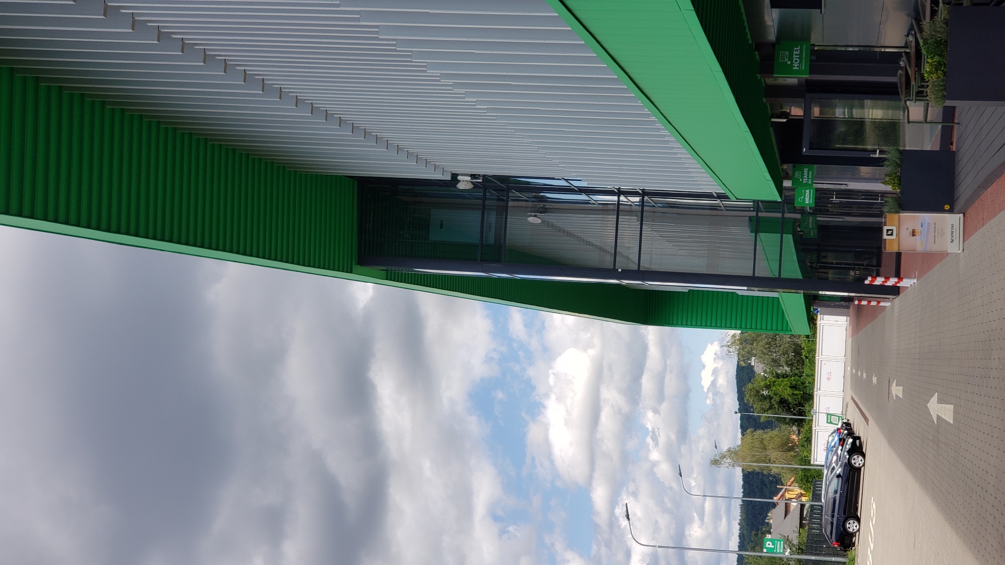 The image shows a modern building with a green roof and silver vertical siding, located in Prešov, Slovakia. The building has large glass windows and entrances. In front of the building, there is a paved walkway with white arrows indicating traffic flow. Several cars are parked in a lot to the left of the walkway. The sky is cloudy with patches of blue. A sign for "HOTEL" is visible on the building's facade. There are also signs for "MEDIA" and "TEAMS". A Nespresso display is positioned near one of the entrances. The surrounding environment includes trees and houses in the background, with hills beyond.