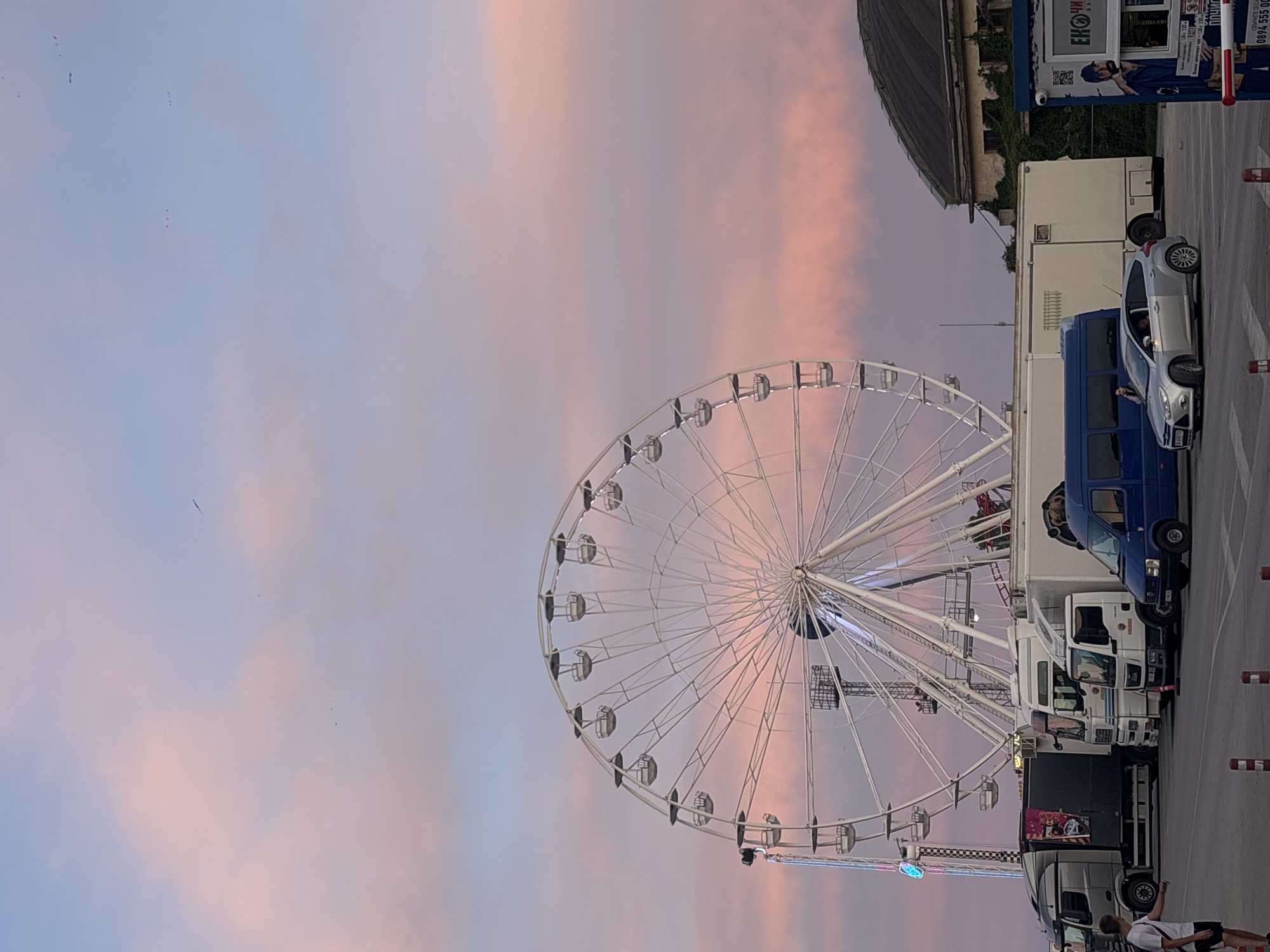 The image depicts a Ferris wheel and several trucks parked in a lot, likely a fairground or event space, with a soft pink and blue sky in the background. The Ferris wheel, a large white structure, dominates the left side of the frame, with its spokes and gondolas clearly visible. To the right of the Ferris wheel, a row of trucks is parked, including a silver semi-truck with a graphic on its side, white box trucks, and a blue van. A silver hatchback car is also parked in the lot, and a person is standing near it. In the background, a weathered building with a dark, curved roof can be seen. The sky exhibits a sunset or sunrise glow with wispy clouds in shades of pink and purple. The overall impression is one of a calm, possibly late evening or early morning scene. The context of Varna, Bulgaria, would suggest this is a public area, possibly a festival or amusement park located within the city.