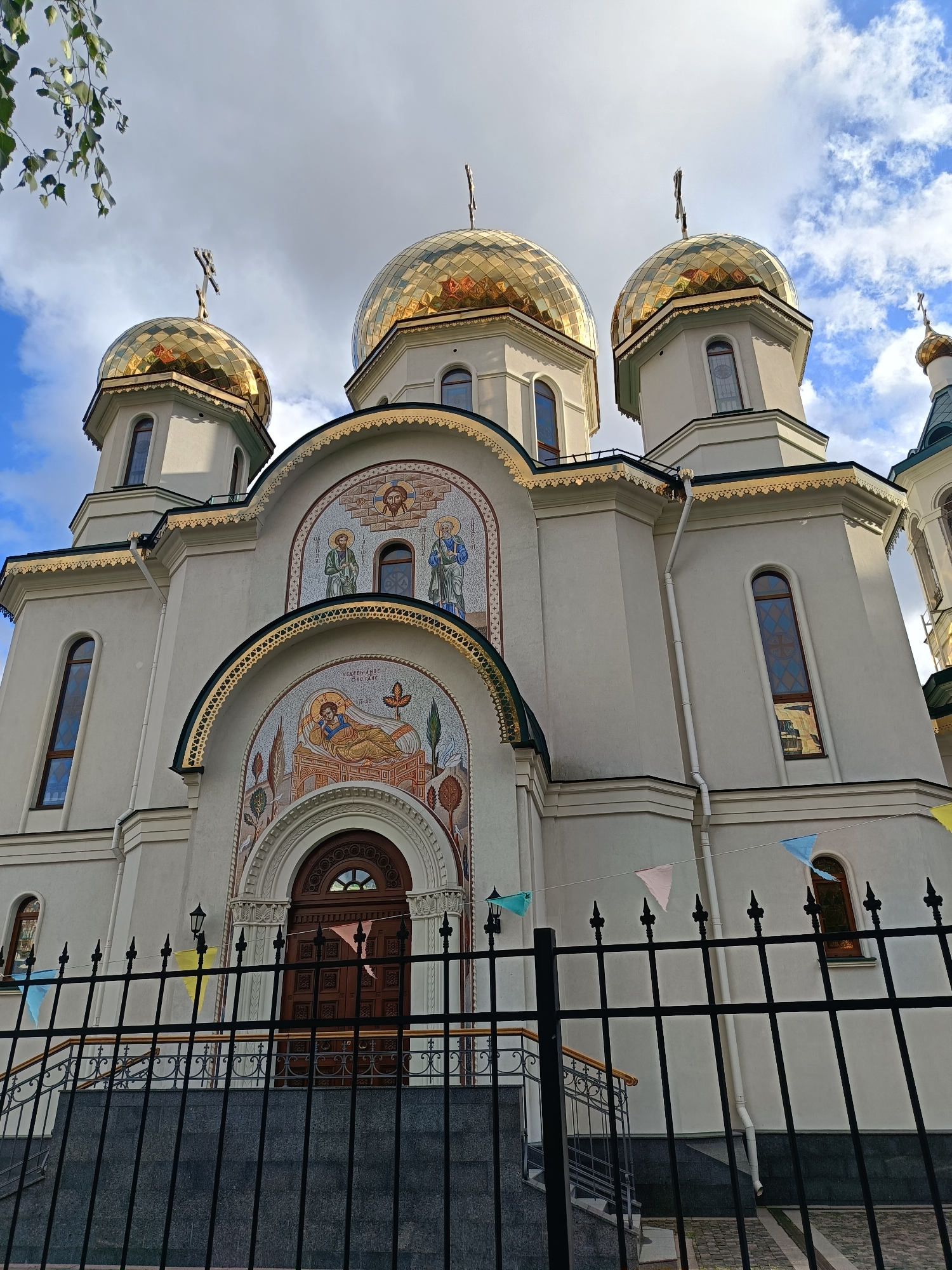The image shows the exterior of a church with multiple golden domes, characteristic of Orthodox Christian architecture. The church is a light color, possibly white or cream, and features arched windows. There are mosaics above the entrance depicting religious figures. A black metal fence is in the foreground, with a stone staircase leading up to the entrance. Small, colorful triangular flags are strung across the fence. The sky is partly cloudy. The style of the church and its adornments suggest it is located in Saint Petersburg, Russia, a city with many Orthodox churches. However, without specific identifying markers or landmarks, the exact location within Saint Petersburg cannot be determined from the image alone.