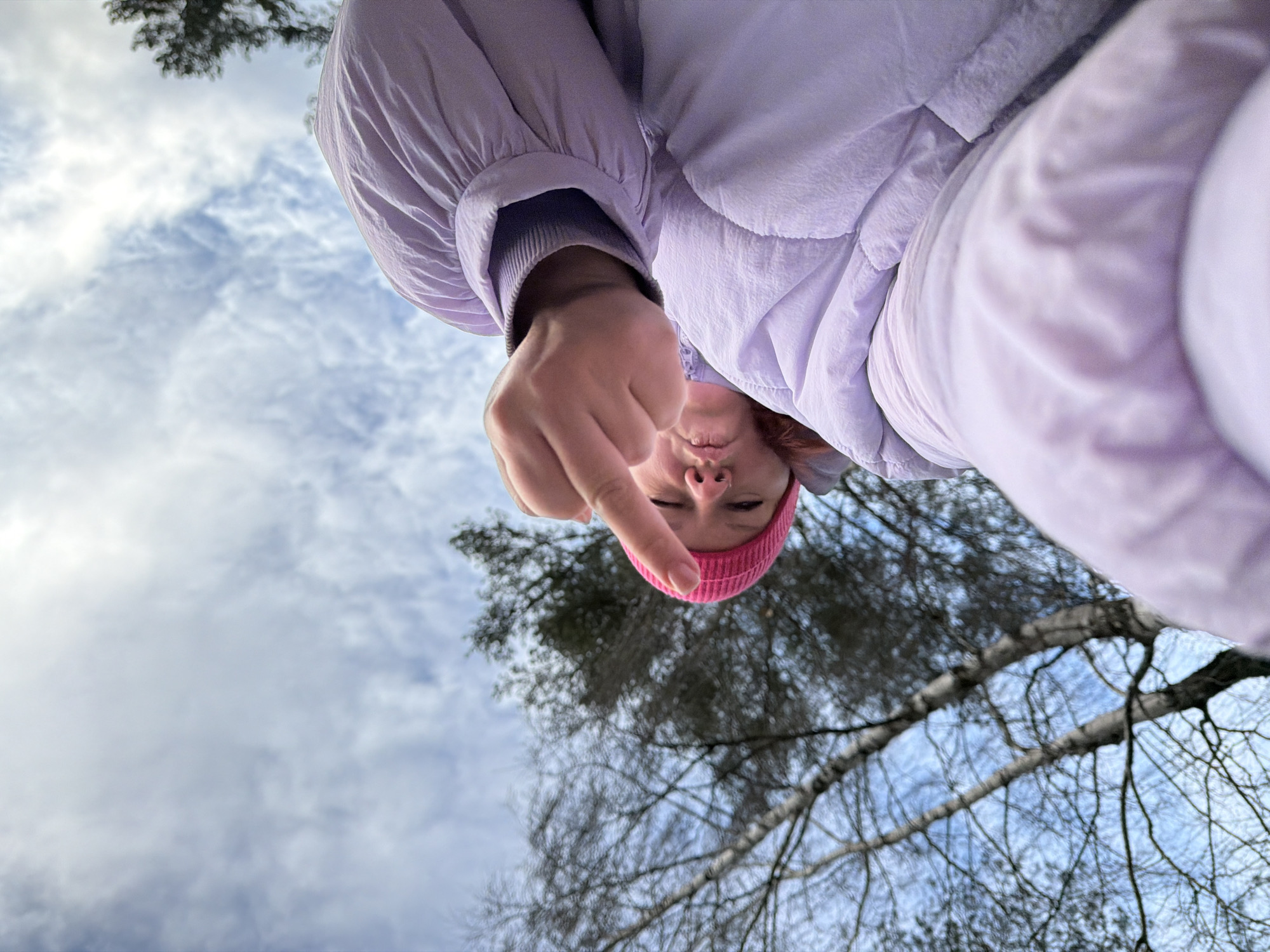 A woman wearing a pink beanie and light purple puffy jacket is captured in a low-angle selfie. She extends her middle finger towards the camera. The background includes bare tree branches against a cloudy sky. The location is presumed to be in or around the city of Chaikovsky, Russia, based on the query.