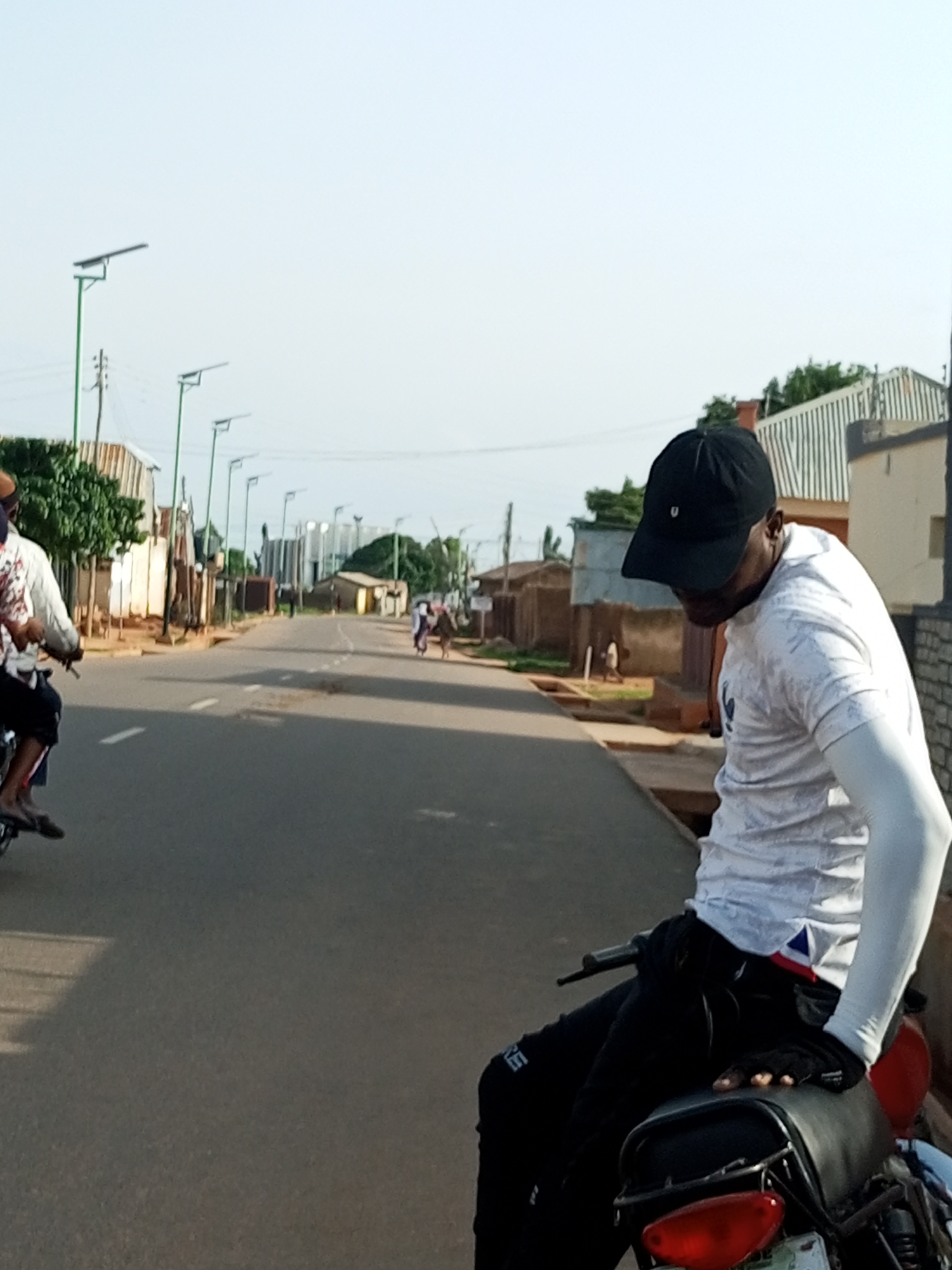 In Tumfure, Nigeria, a man wearing a white t-shirt with a patterned design and a black baseball cap is seated on a motorcycle. He has on black pants and fingerless gloves. The motorcycle is parked on an asphalt road lined with buildings on both sides. In the background, there are streetlights, other motorcycles, and people walking. The sky is clear and bright, indicating daytime.
