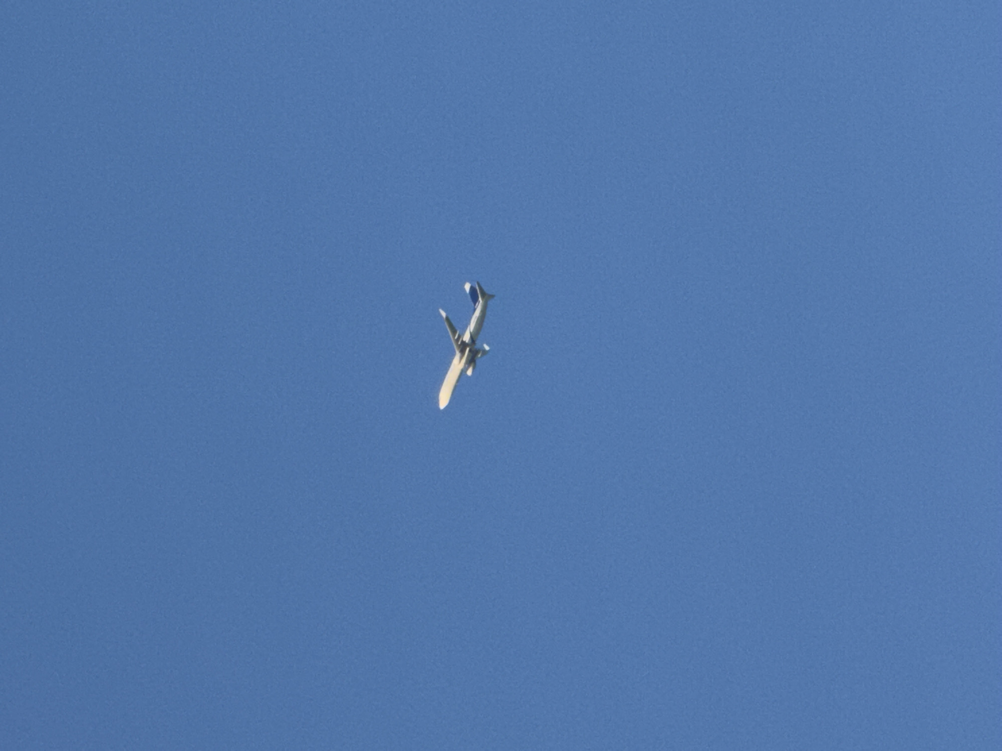 An airplane is captured mid-flight against a clear blue sky. The aircraft is white with blue accents on its tail. The image is taken from below, looking up at the plane. The specific location in Tbilisi, Georgia, cannot be determined from the provided image.