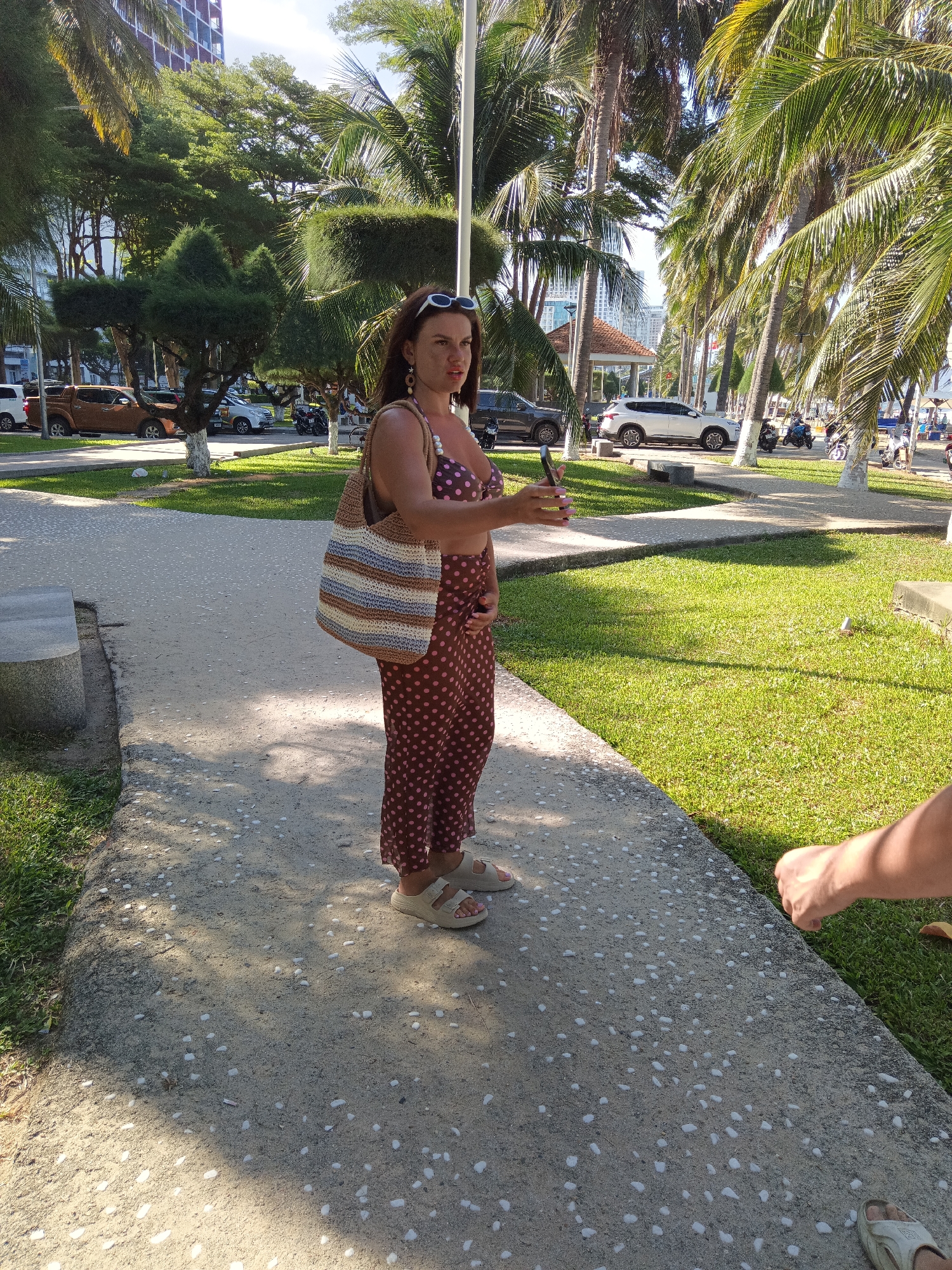 A woman stands on a paved walkway in a park-like setting with palm trees and manicured bushes. She is wearing a brown polka-dot bikini top and a matching polka-dot skirt, and is carrying a woven bag over her shoulder. She holds a cell phone in her right hand. A portion of another person's arm and hand is visible in the lower right corner of the image, reaching towards the woman. In the background, cars and motorcycles are parked, and a few people are walking. A building with a red top is visible in the distance. This scene is consistent with the beachfront areas of Nha Trang, Vietnam, which often feature palm-lined walkways, grassy areas, and parking for vehicles.
