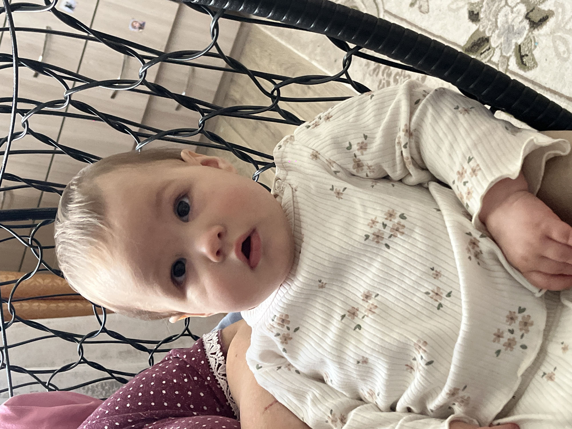 An upward-angled, close-up photograph captures a baby with light-colored hair and blue eyes, looking directly at the camera with a curious expression and slightly open mouth. The baby is wearing a light-colored, ribbed long-sleeved top adorned with small, delicate floral patterns in muted brown and green tones. A portion of a person's maroon clothing with white polka dots and lace trim is visible on the lower left, suggesting the baby is being held or sitting on someone's lap.
The scene is indoors, likely a home in Novopokrovskaya, Russia. A prominent black, interwoven wire structure, possibly part of a decorative chair or swing, occupies the left side and foreground, framing the baby. In the background, to the right, a light-colored patterned rug or carpet is visible. Above the black structure, there appears to be a light-colored ceiling or wall, with faint lines and two small, indistinct framed pictures or decorations. The lighting suggests it is daytime. No visible text is present in the image.