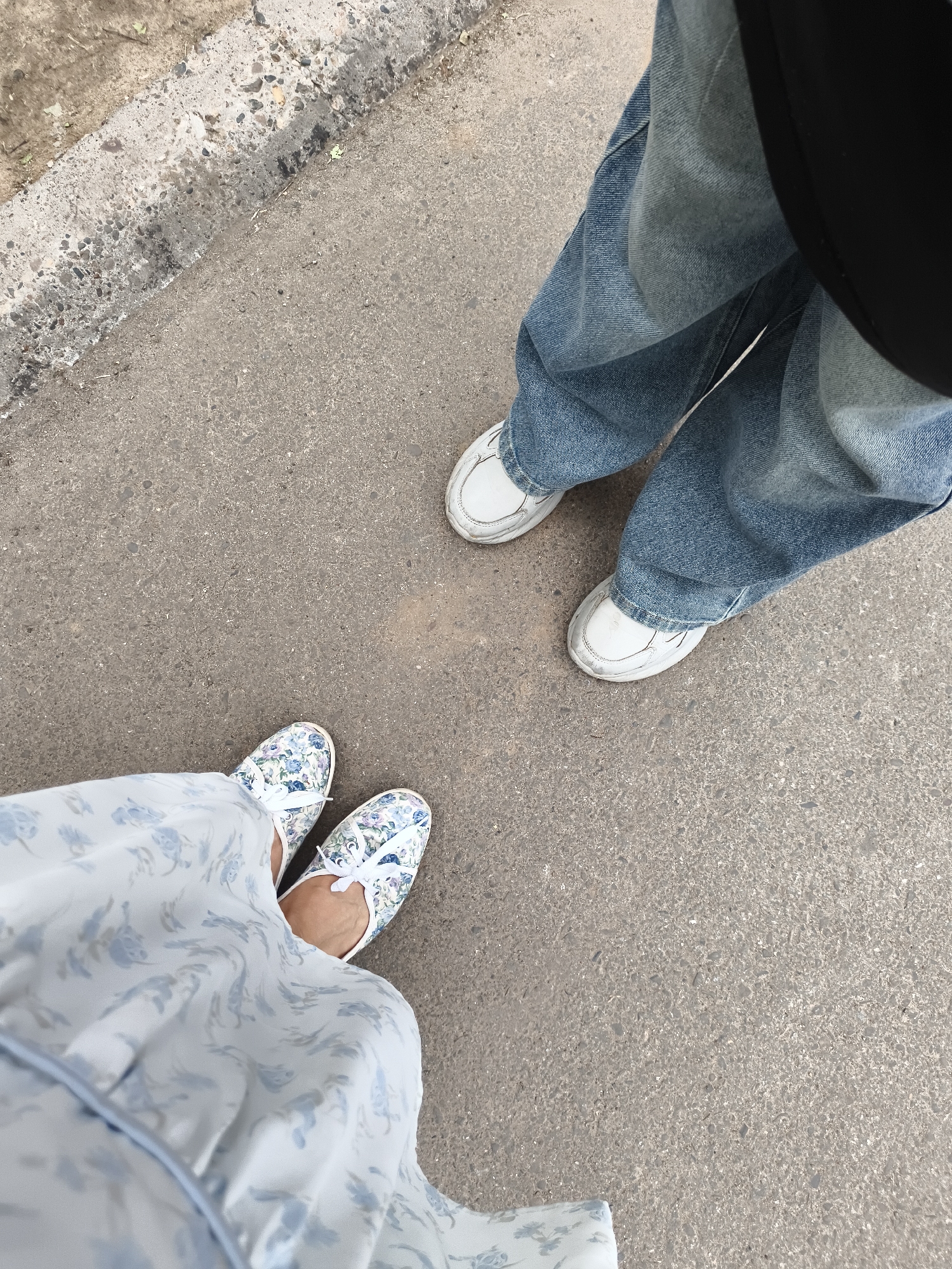Two pairs of feet are visible on an asphalt surface. One pair is adorned with floral-patterned slip-on shoes and a light blue dress with a similar floral print. The other pair is wearing white sneakers and blue denim jeans. The scene suggests a casual outing. The lighting indicates it is daytime, likely sunny, given the lack of shadows. The asphalt surface and the concrete curb in the background suggest an outdoor urban or suburban setting. No text is visible.