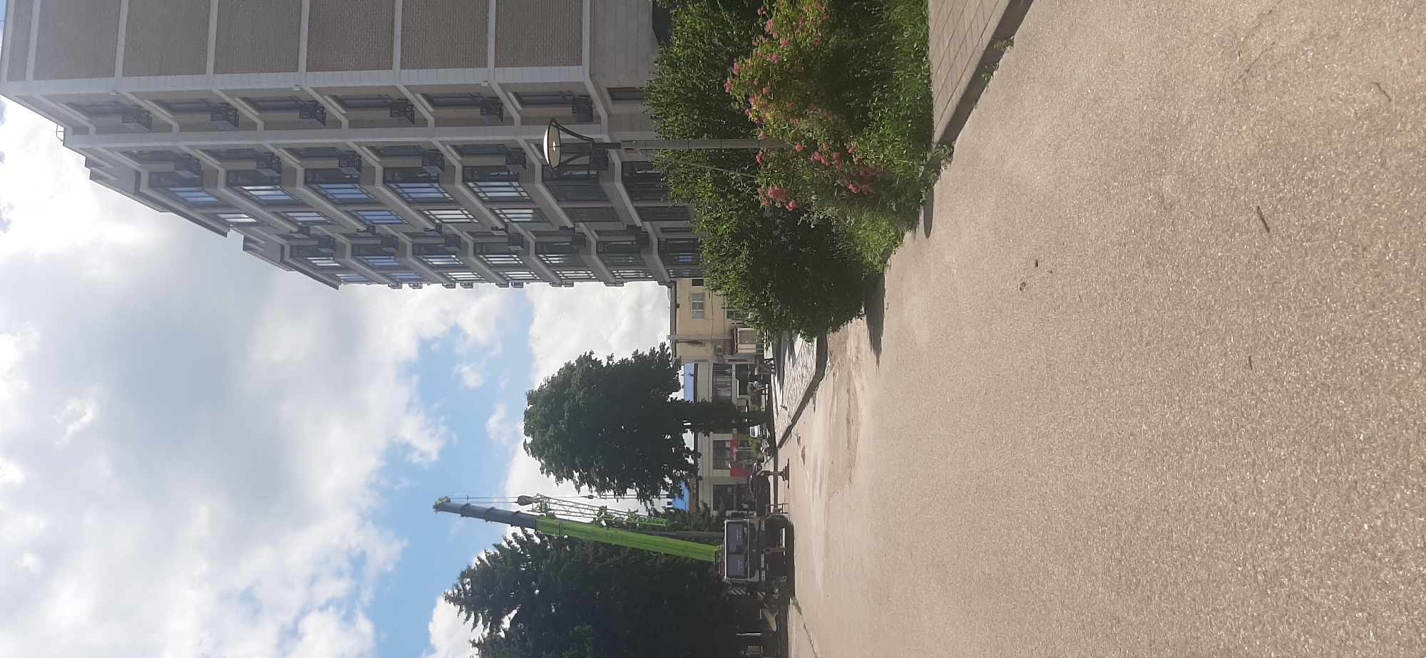 A construction crane is visible on the left side of the frame, with its arm extended upwards. In the background, a multi-story building with numerous windows stands on the right, and other buildings are visible further back. Several trees are present, including a large deciduous tree in the center and evergreen trees on the left. A paved road or walkway extends into the foreground. The sky is partly cloudy. The image depicts a scene in Yessentukskaya, Russia, with evidence of ongoing construction work.