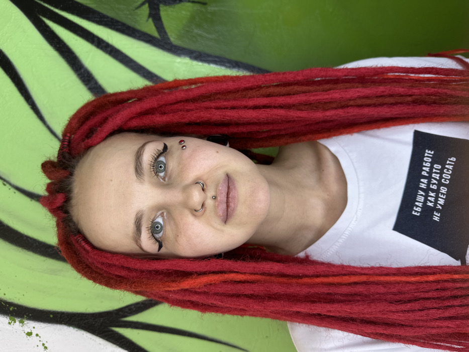 A head-and-shoulders portrait depicts a female subject, estimated to be a young adult, with fair skin, light eyes, and prominent red dreadlocks extending past her shoulders. She has a silver circular barbell piercing in her septum, a silver stud in her philtrum, and two red dermal jewel piercings on her left cheekbone. Her ears feature dark-colored earlobe plugs. Her eyes are adorned with winged eyeliner and mascara. She wears a white t-shirt with a black rectangular graphic on the front, displaying white Russian text that translates to "I WORK MY ASS OFF AS IF I DON'T KNOW HOW TO SUCK." The subject maintains a neutral facial expression, looking directly at the camera. The background consists of a bright lime green wall with black graffiti-style outlines and white paint splatters. This scene is located in the city of Saratov, Russia.