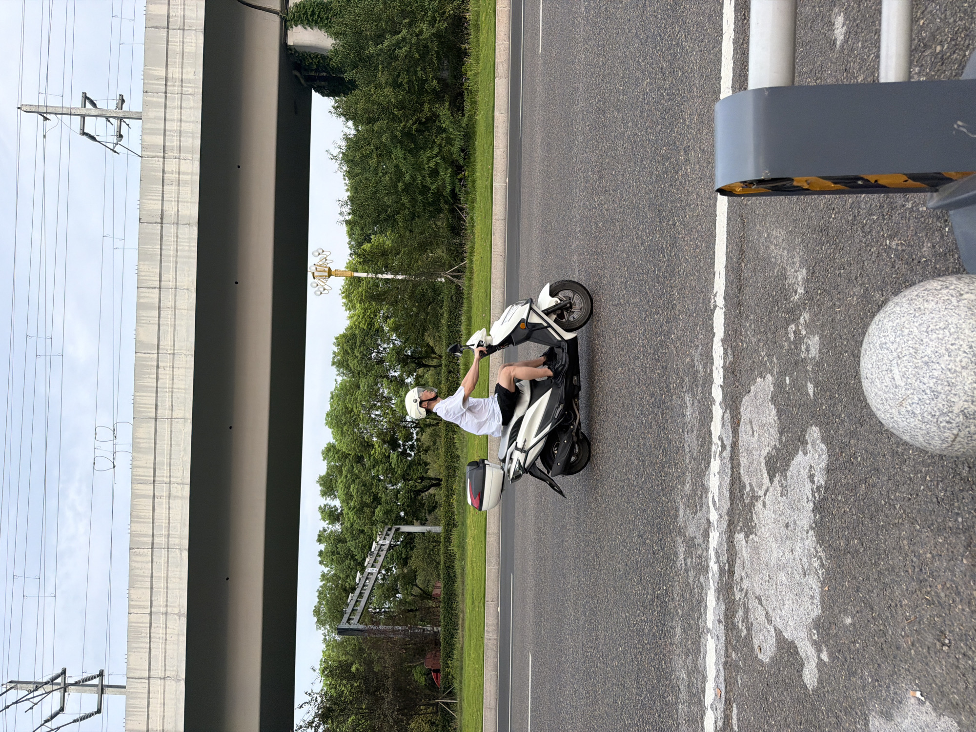 A person wearing a white shirt, black shorts, and a white helmet is riding a white and black scooter on an asphalt road. The scooter has a white storage box on the back. The person is looking forward. The road has a white line marking its edge. In the background, there are trees, a street lamp, and an elevated overpass with power lines running overhead. The sky is overcast. The scene appears to be in an urban or suburban area in Jiangjin, China.