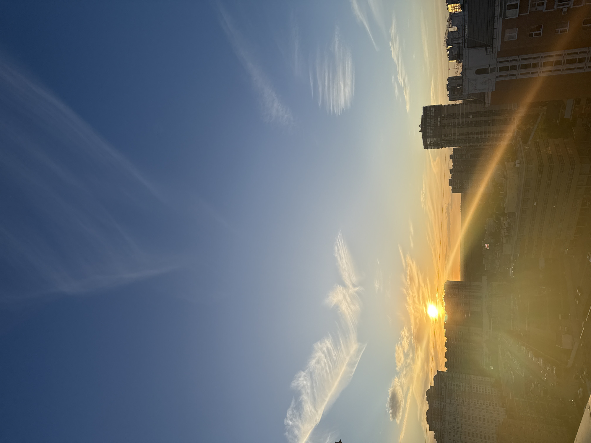 The image captures a sunset over the city of Khimki, Russia. Tall residential buildings dominate the skyline, their structures silhouetted against a vibrant sky. The sun, a bright orb, is positioned near the horizon, casting long rays of light that illuminate the scene with a warm, golden glow. Wispy clouds are scattered across the blue expanse, some tinged with the colors of the sunset. Below the buildings, a street is visible, lined with parked cars, suggesting an urban environment. The overall impression is one of a peaceful evening in a bustling city.