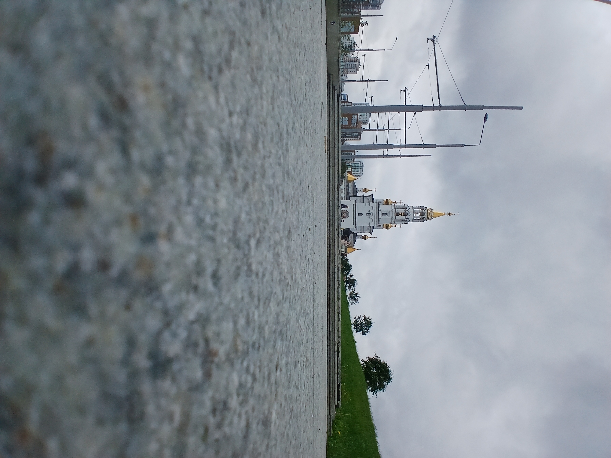 The image shows a low-angle view of a stone surface in the foreground, with a church and urban landscape in the background. The church is white with golden domes and crosses, and appears to be the Church of the Assumption of the Most Holy Theotokos in Yekaterinburg, Russia. The church is surrounded by trees and a grassy hill on the right. In the mid-ground, there are tram lines and poles, along with several multi-story buildings. The sky is overcast and gray, suggesting a cloudy day. There are a few figures visible near the church, but they are small and indistinct. The overall scene depicts a typical urban environment with a prominent religious building.