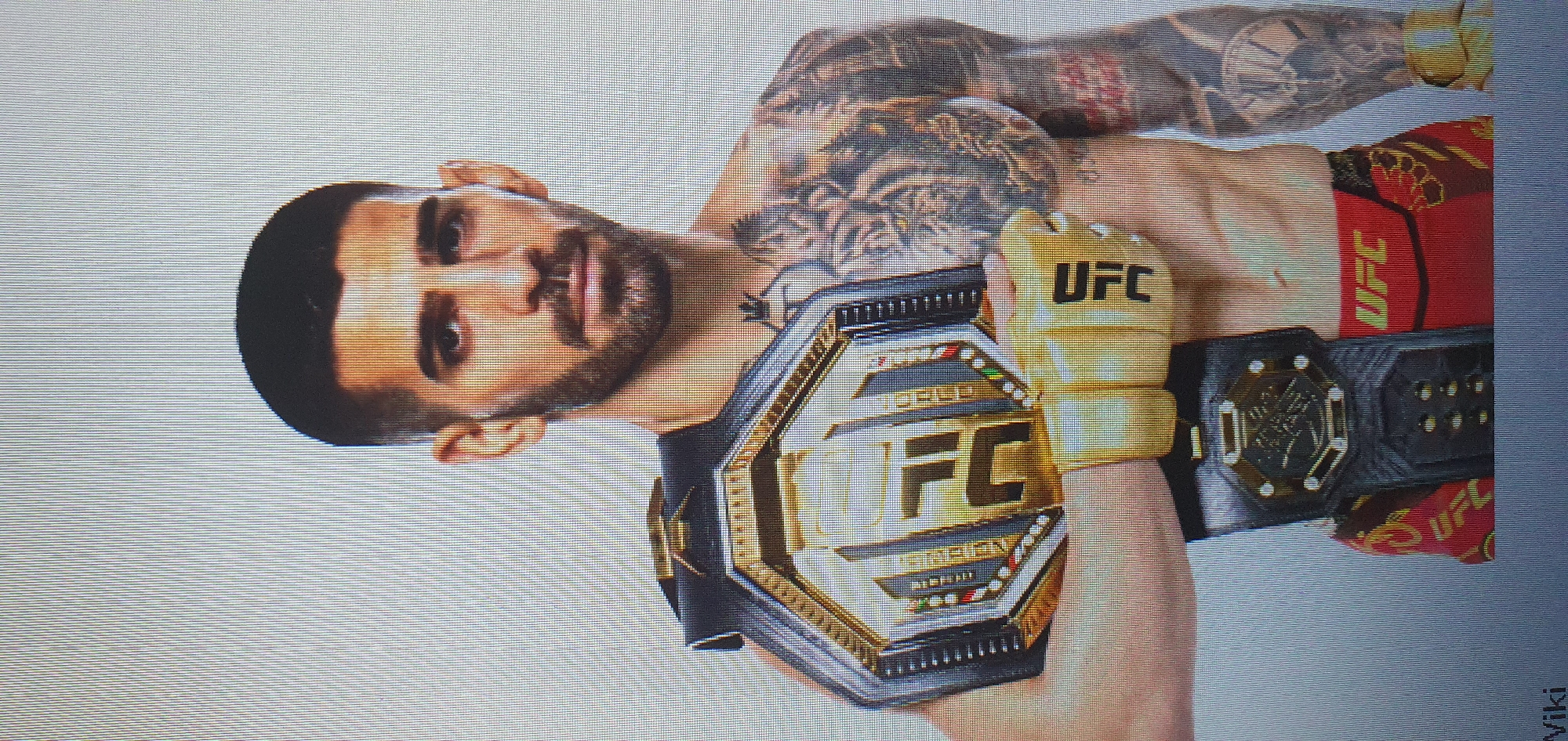This image is a studio portrait of a male UFC fighter. He is wearing a championship belt across his chest and gold UFC gloves on his hands. The fighter has short dark hair, a beard, and tattoos on his chest and arms. He is wearing red UFC shorts with gold accents. The background is a plain, light gray. There is no indication of the city of Bat Yam, Israel, in the image.
