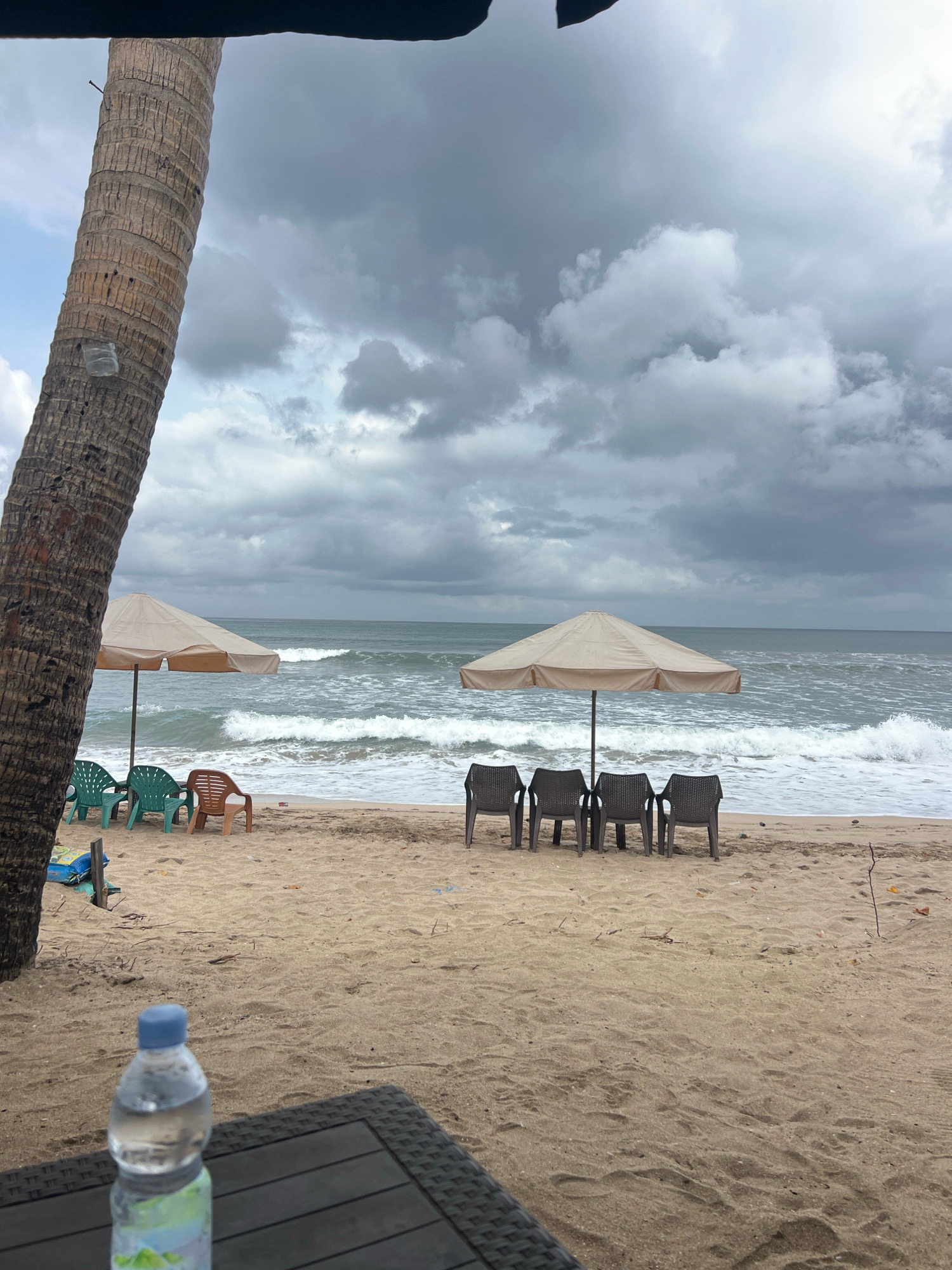 The image depicts a beach scene in Kuta, Indonesia, under a cloudy sky. In the foreground, a dark, textured table with a partially visible bottle of water sits on the sand. The beach itself is sandy, with scattered debris. Several plastic chairs, some green and one orange, are arranged near a light-colored beach umbrella. Further down the beach, a row of four dark plastic chairs is positioned under another similar umbrella. The ocean is visible in the background, with moderate waves breaking on the shore. A thick palm tree trunk is prominently featured on the left side of the image. There are no people visible in the scene. The overall atmosphere is overcast and slightly moody, suggesting a change in weather.