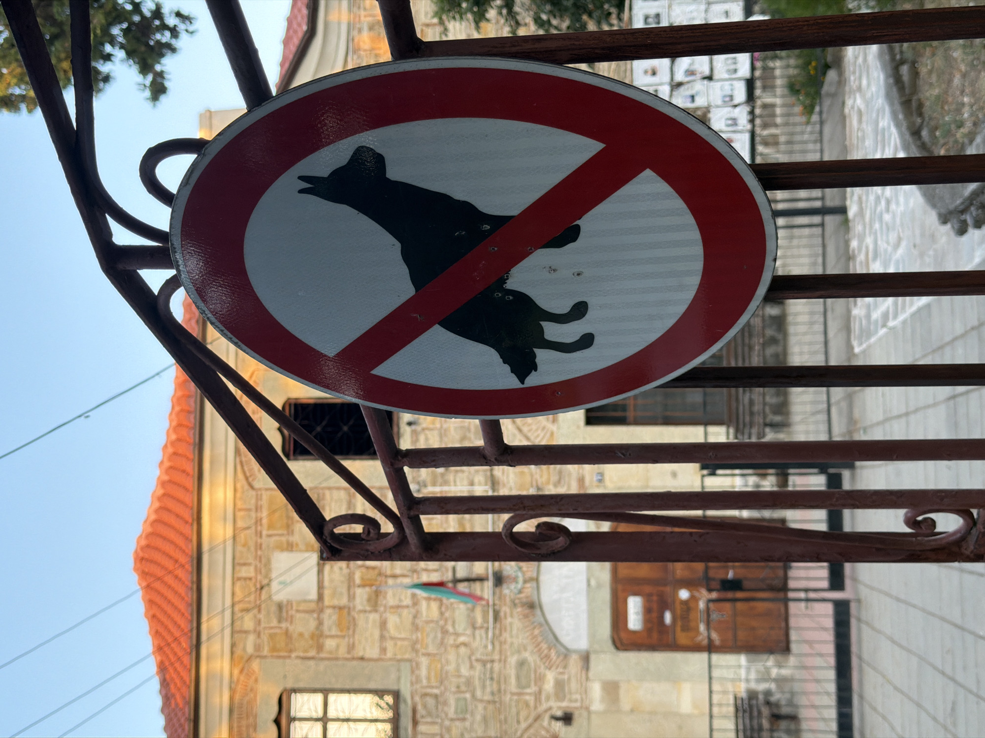 A no dogs allowed sign is visible on a rusted metal fence in front of a stone building. The building has a red tiled roof and a Bulgarian flag displayed. The sign is circular with a red border and a red diagonal line crossing a silhouette of a dog. The background shows a paved walkway and some greenery.