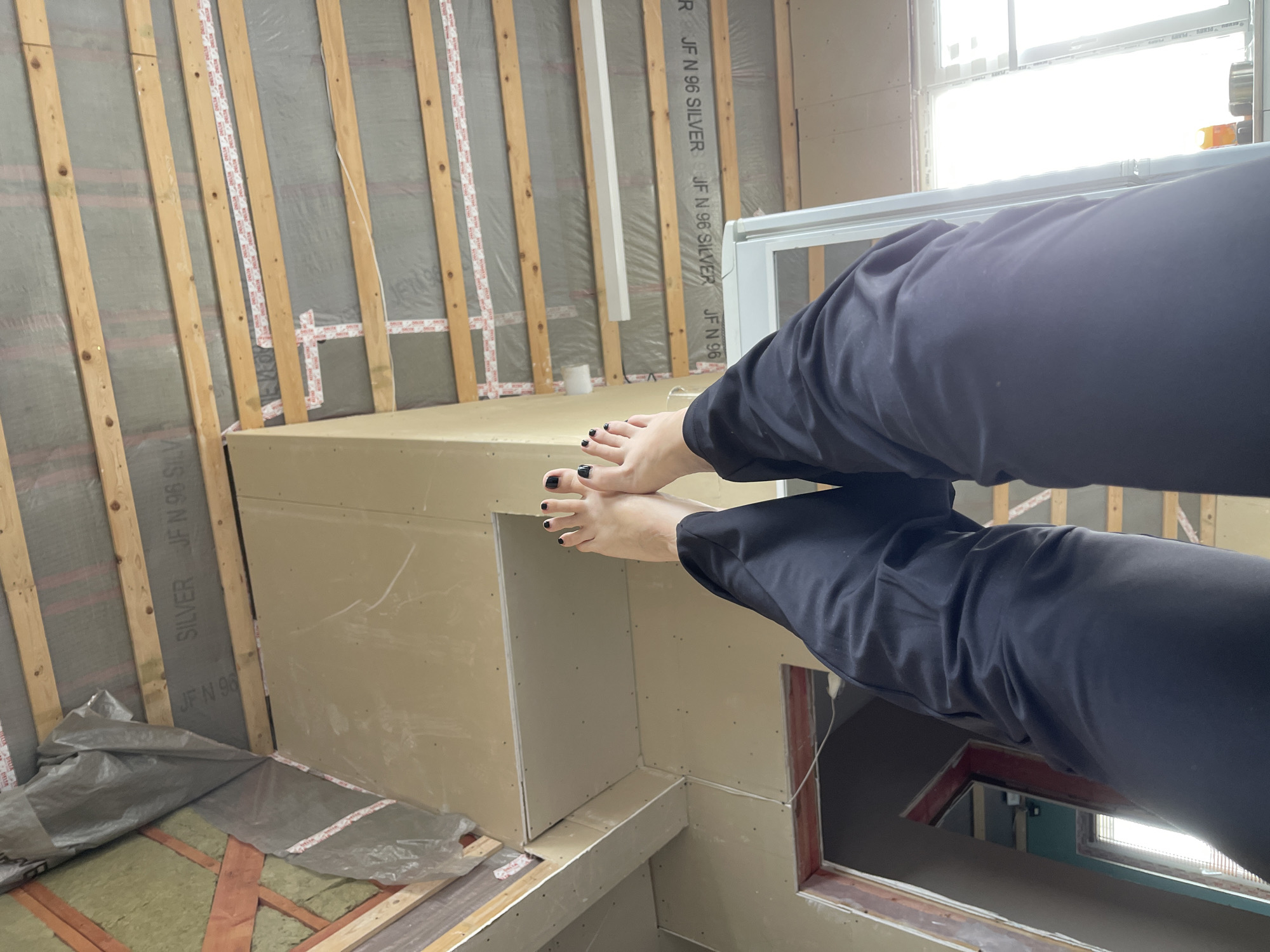 The image appears to be taken inside a room under construction. The interior is raw, with exposed wooden framing and insulation materials visible on the walls. A structure made of drywall or similar material sits in the foreground, possibly a built-in unit or cabinet. A window allows natural light to enter the space. A pair of legs wearing dark pants and feet with painted toenails are positioned centrally in the frame, obscuring some of the background. The location of the room appears to be in Мосметаллоконструкция Онт, Russia.