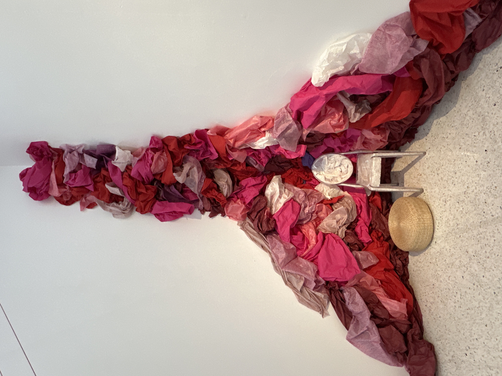 The image shows an art installation on a white wall next to a speckled floor. The installation is composed of multiple pieces of colored tissue paper in shades of red, pink, and white. The paper is arranged to resemble a spreading, irregular shape, emanating from a central point near the wall/floor junction. A small metal shelf-like structure with two round objects (one white and one tan) is affixed to the wall amidst the tissue paper. The scene is indoors. The image does not contain sufficient information to identify the city of Banan, China.