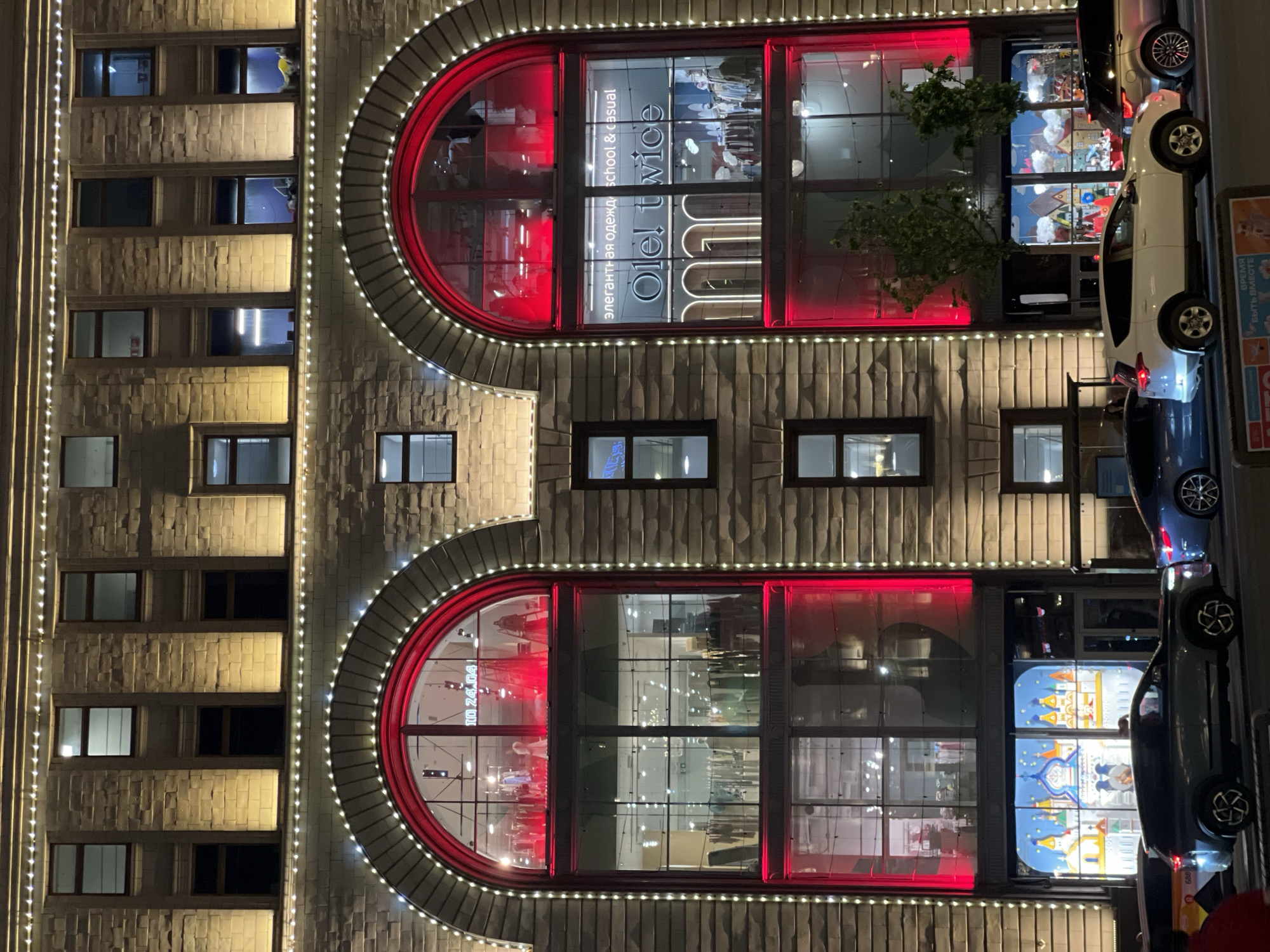 Here is a forensic description of the image.
The image shows the facade of a building, potentially in Moscow, Russia, at night. The building has several windows, including two large arched windows outlined with decorative lights. Red illumination highlights the architectural features around the arched windows. A sign within one window reads "Ole! twice" and includes text in Russian that translates to "Elegant clothing school & casual." The other arched window features displays or decorations. To the right of the building, vehicles are parked along the street. The text "ВРЕМЯ БЫТЬ ВМЕСТЕ" (Time to be Together) appears on a sign near the parked cars. A number appears in the bottom corner of the photo that reads "688."